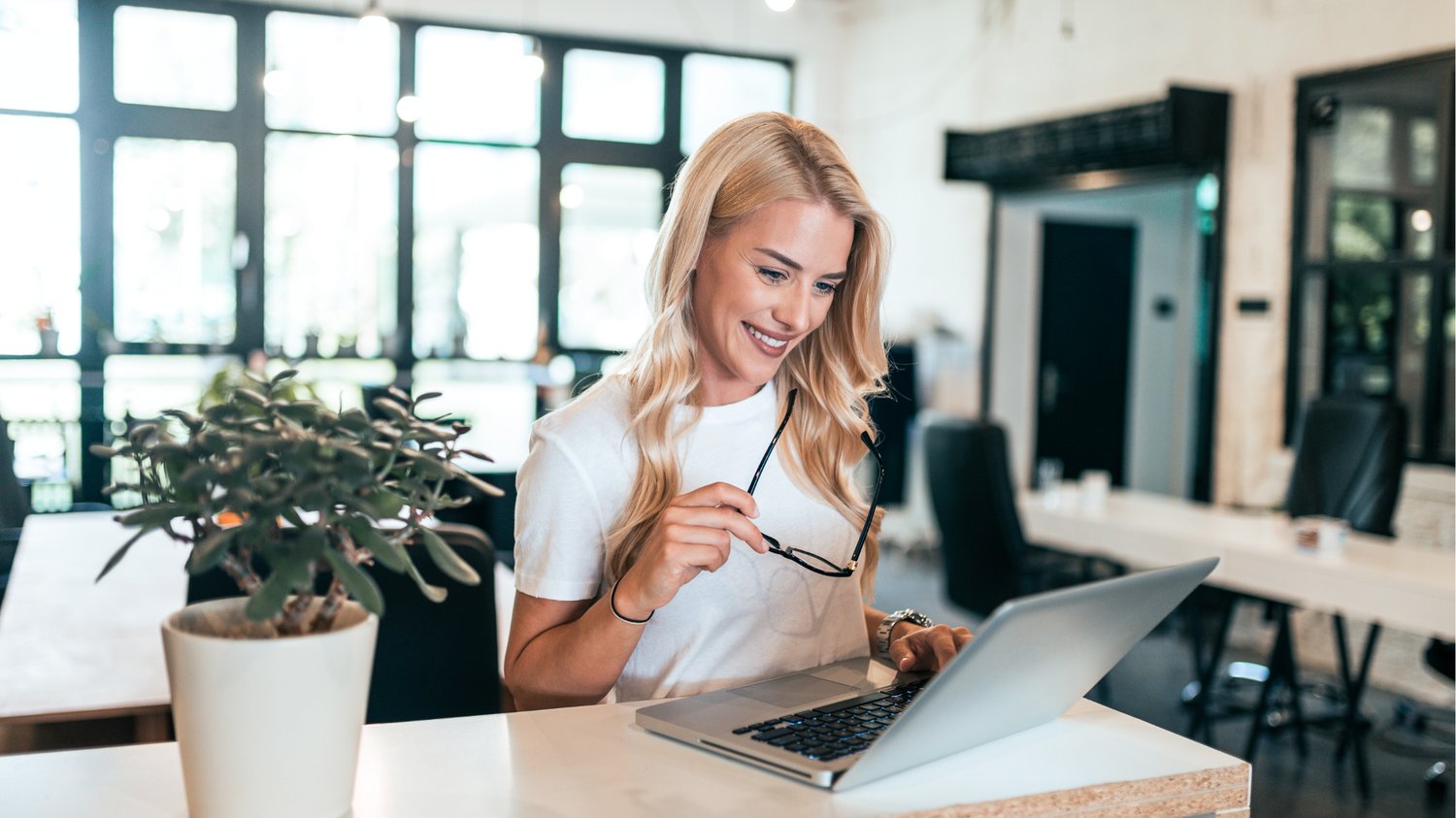 Smiling health coach working on a laptop in a bright, modern workspace, holding glasses and feeling confident—illustrating stress-free content planning with a simple marketing framework.