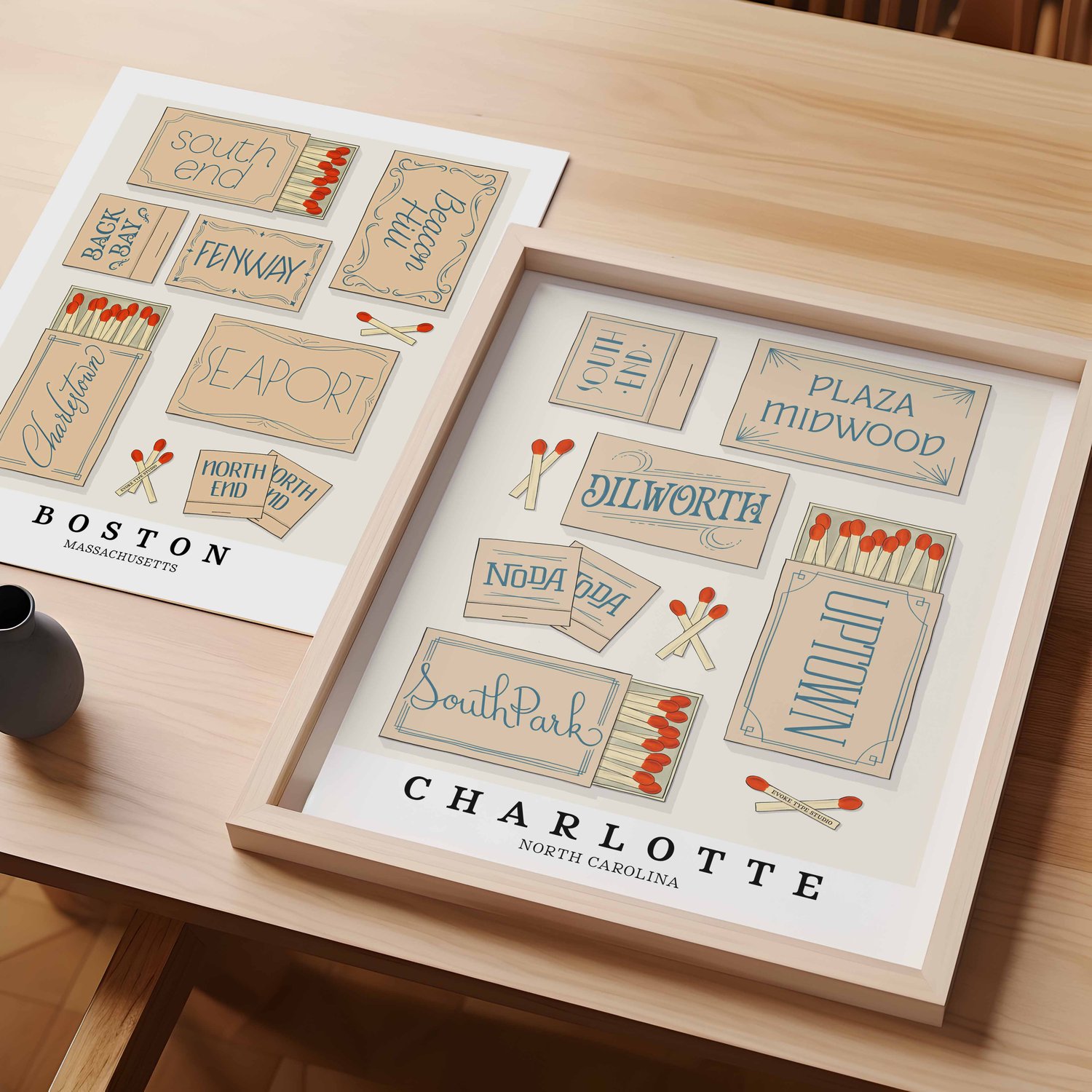 A table with an unframed artwork of Boston matchbox art and a framed artwork of Charlotte matchbox art