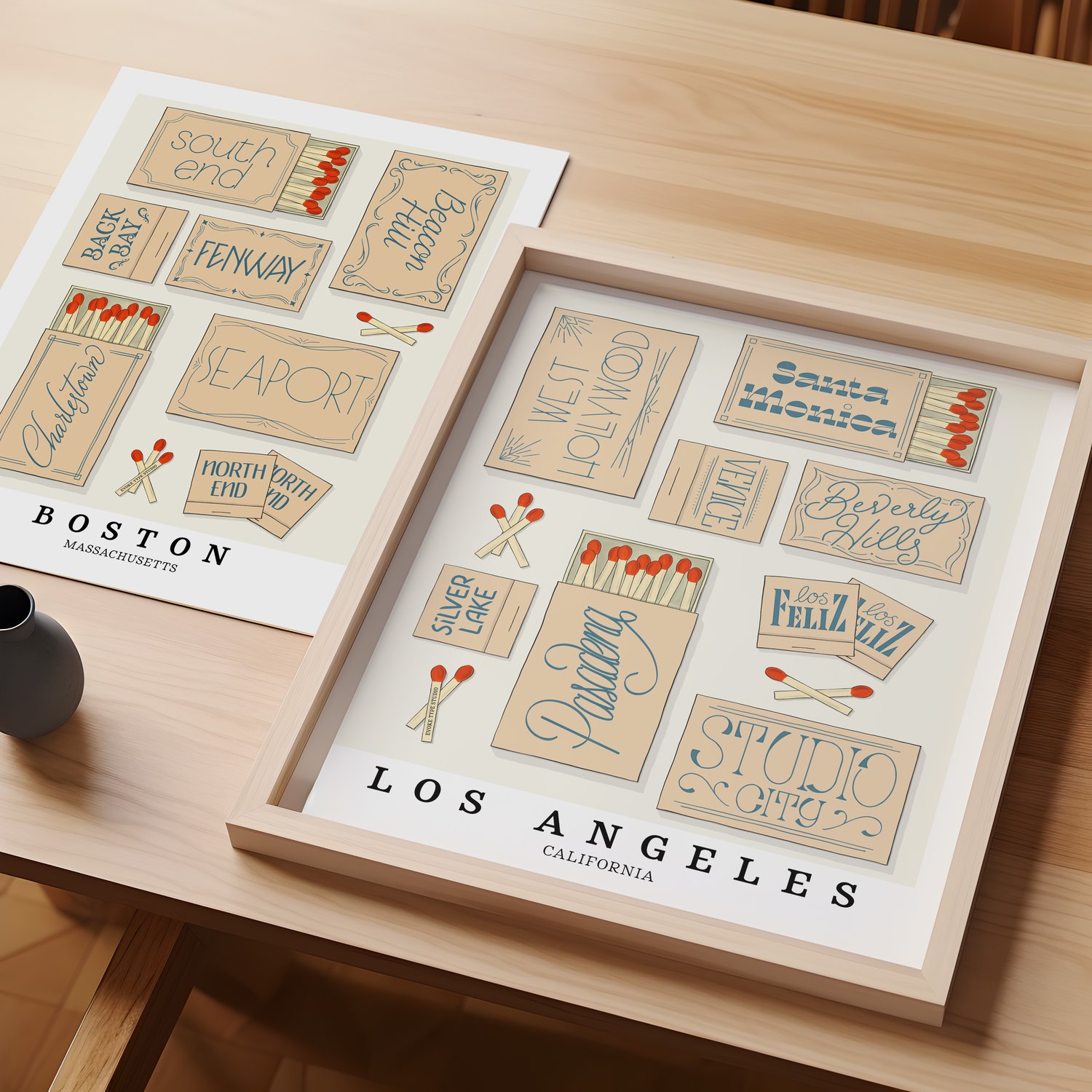 A table with an unframed artwork of Boston matchbox art and a framed artwork of Los Angeles matchbox art