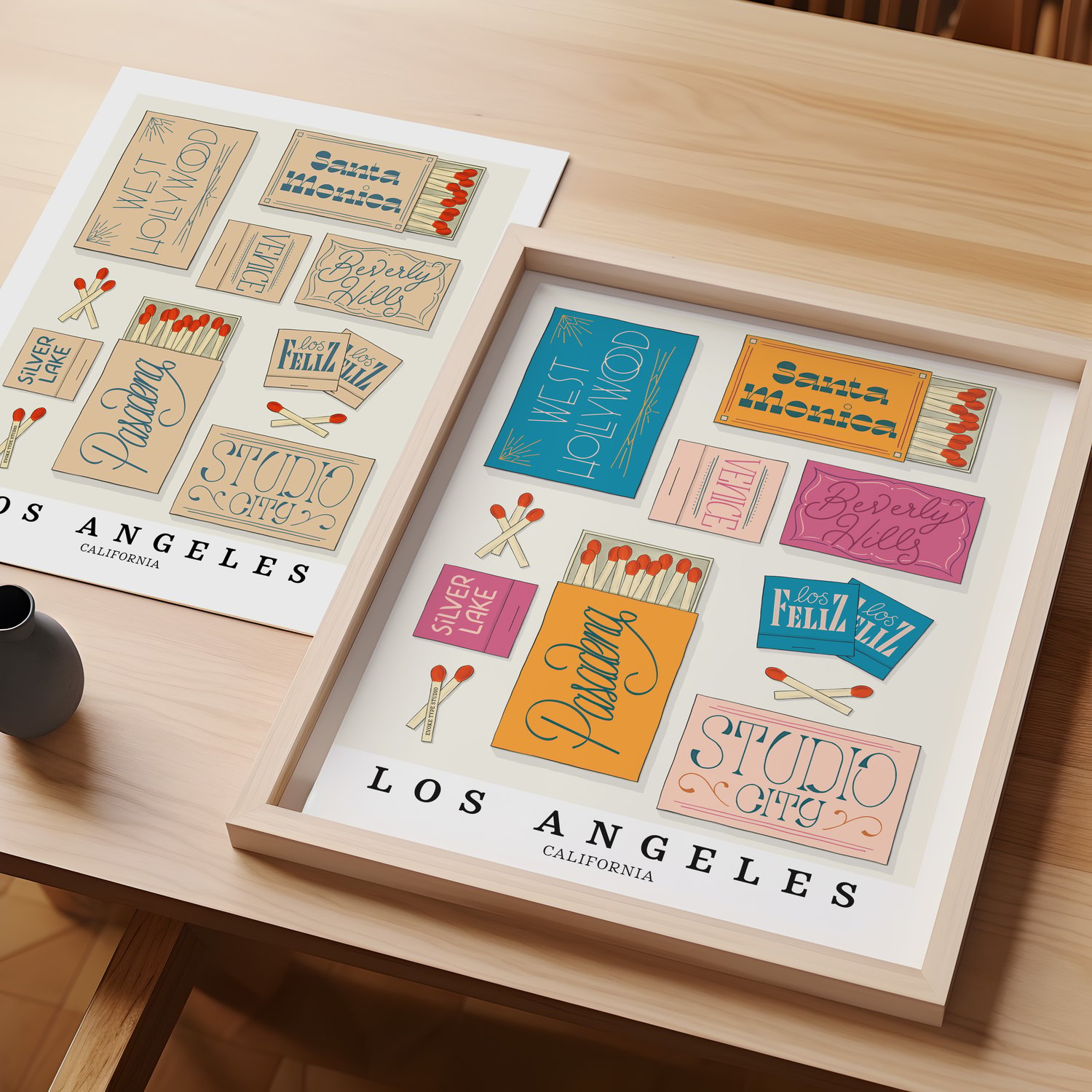 A table with an unframed artwork of Los Angeles matchbox art and a framed artwork of Los Angeles matchbox art