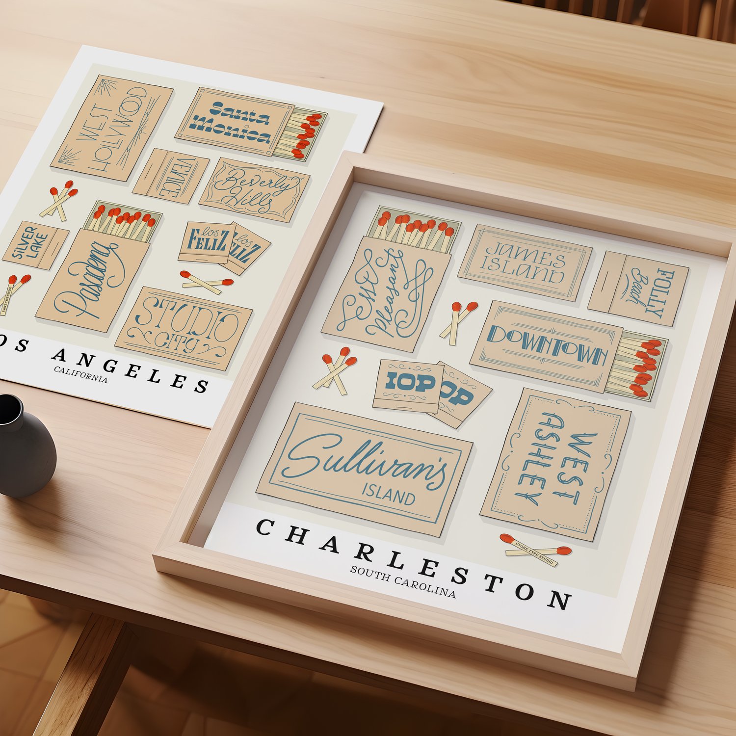 A table with an unframed artwork of Los Angeles matchbox art and a framed artwork of Charleston matchbox art