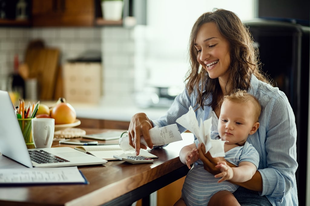 Young working mother with baby using calculator and going through bills while smiling.
