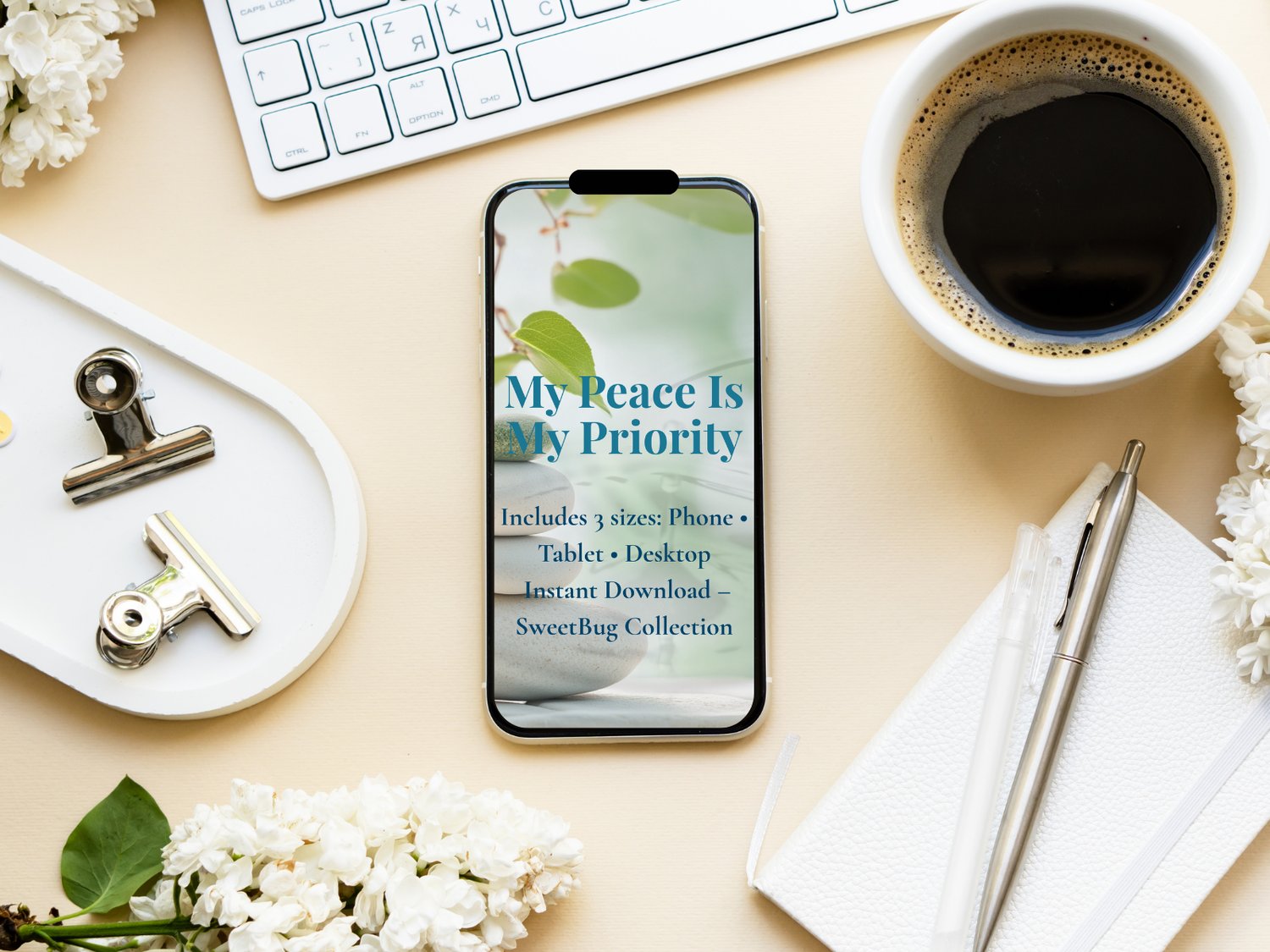 A smartphone displays the affirmation wallpaper "My Peace Is My Priority" on a desk setup. The background includes a peaceful scene of stacked zen stones and green leaves. Surrounding the phone are a cup of black coffee, a keyboard, notepad with a pen, wh