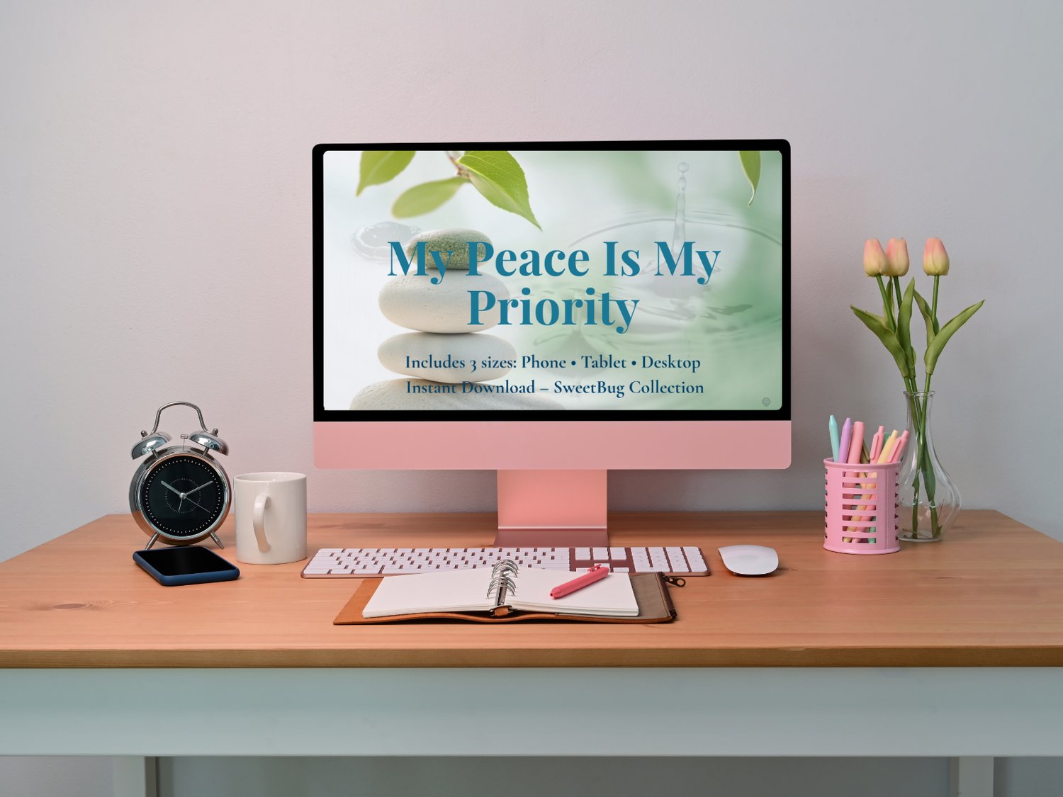 A peaceful workspace featuring a pink desktop computer displaying a calming digital wallpaper titled “My Peace Is My Priority,” with spa stones and green leaves in the background. Desk items include a notebook, clock, tulips, and pens, promoting a tranqui