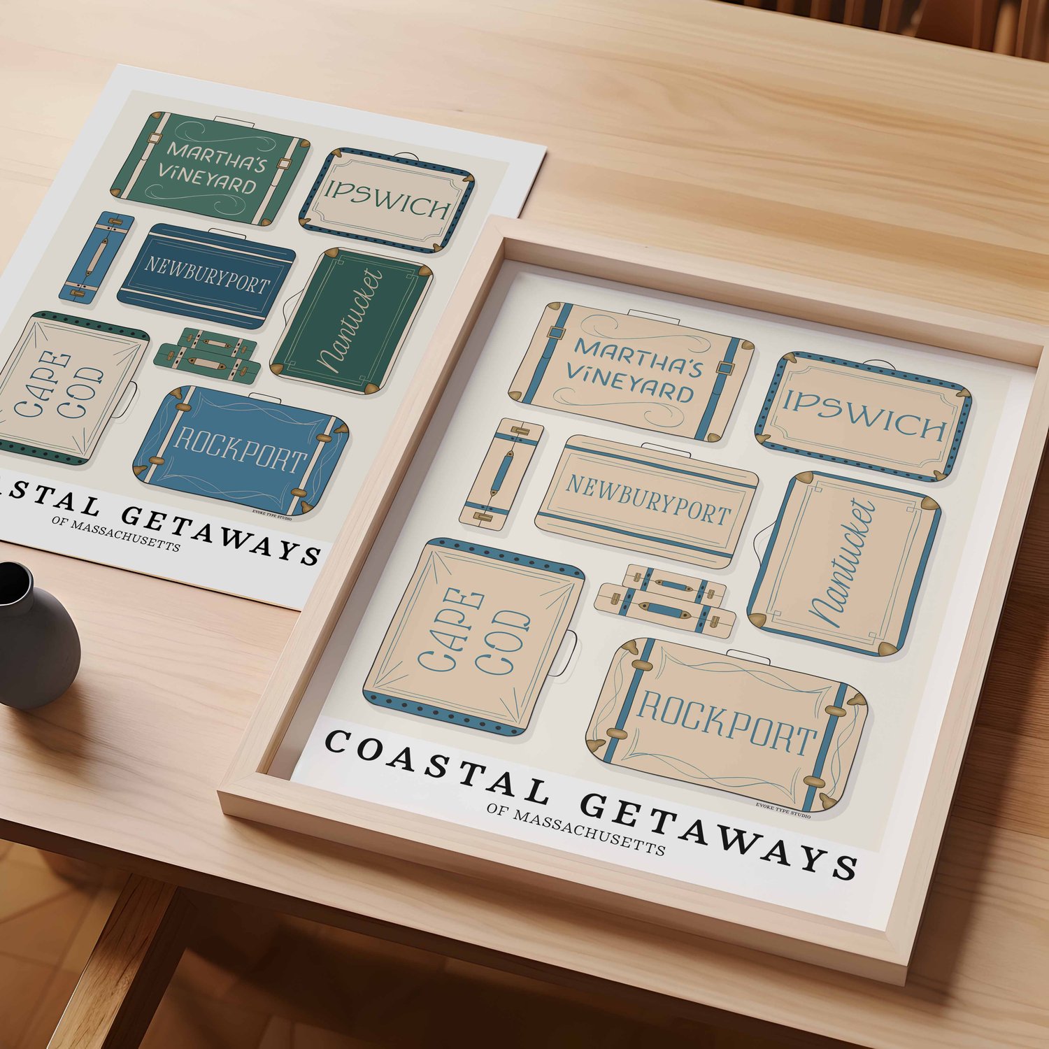 A table with an unframed artwork of Coastal Massachusetts suitcase art and a framed artwork of Coastal Massachusetts suitcase art