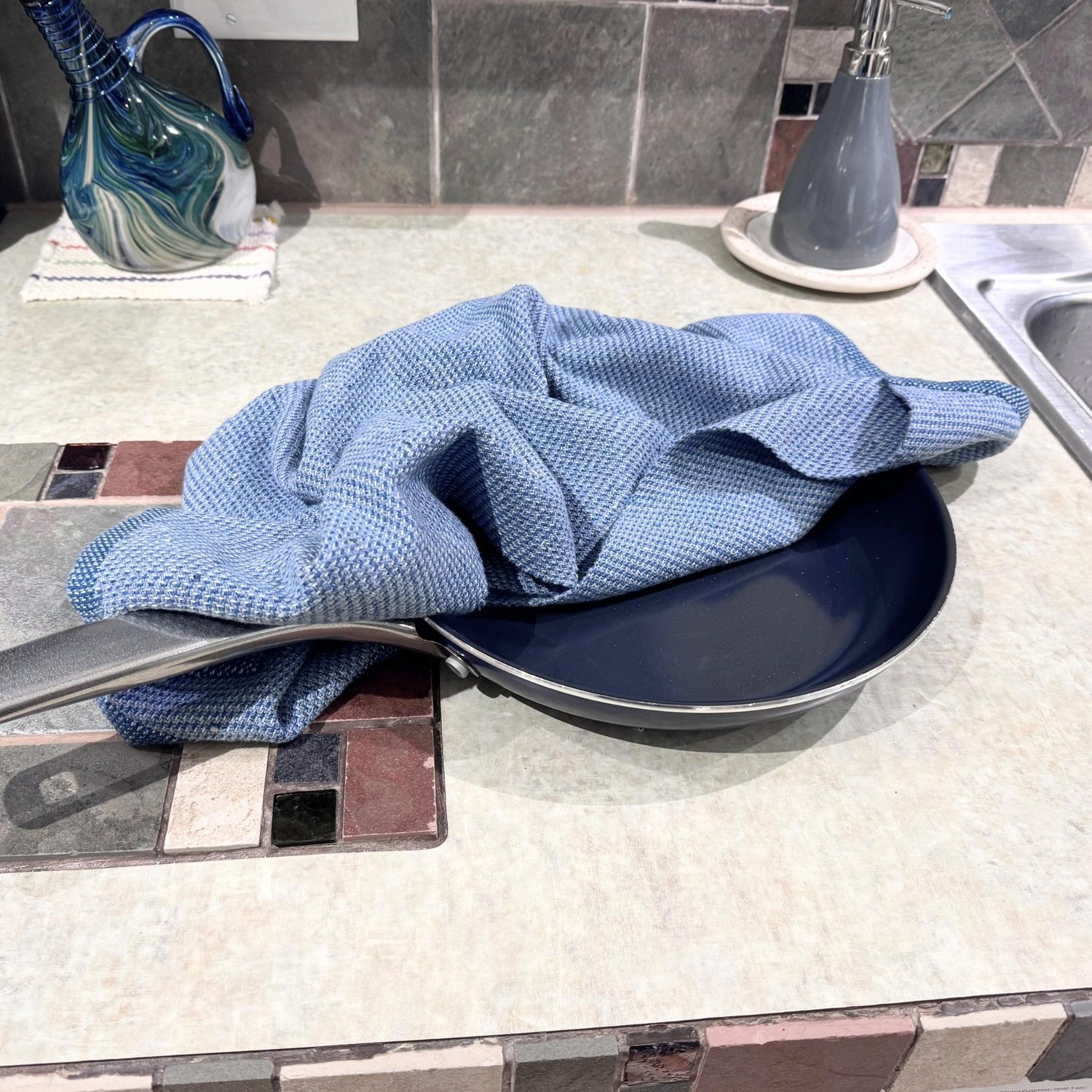 Jeans blue handwoven dish towel displayed with a black frying pan on a counter with brown tile and pale green formica.