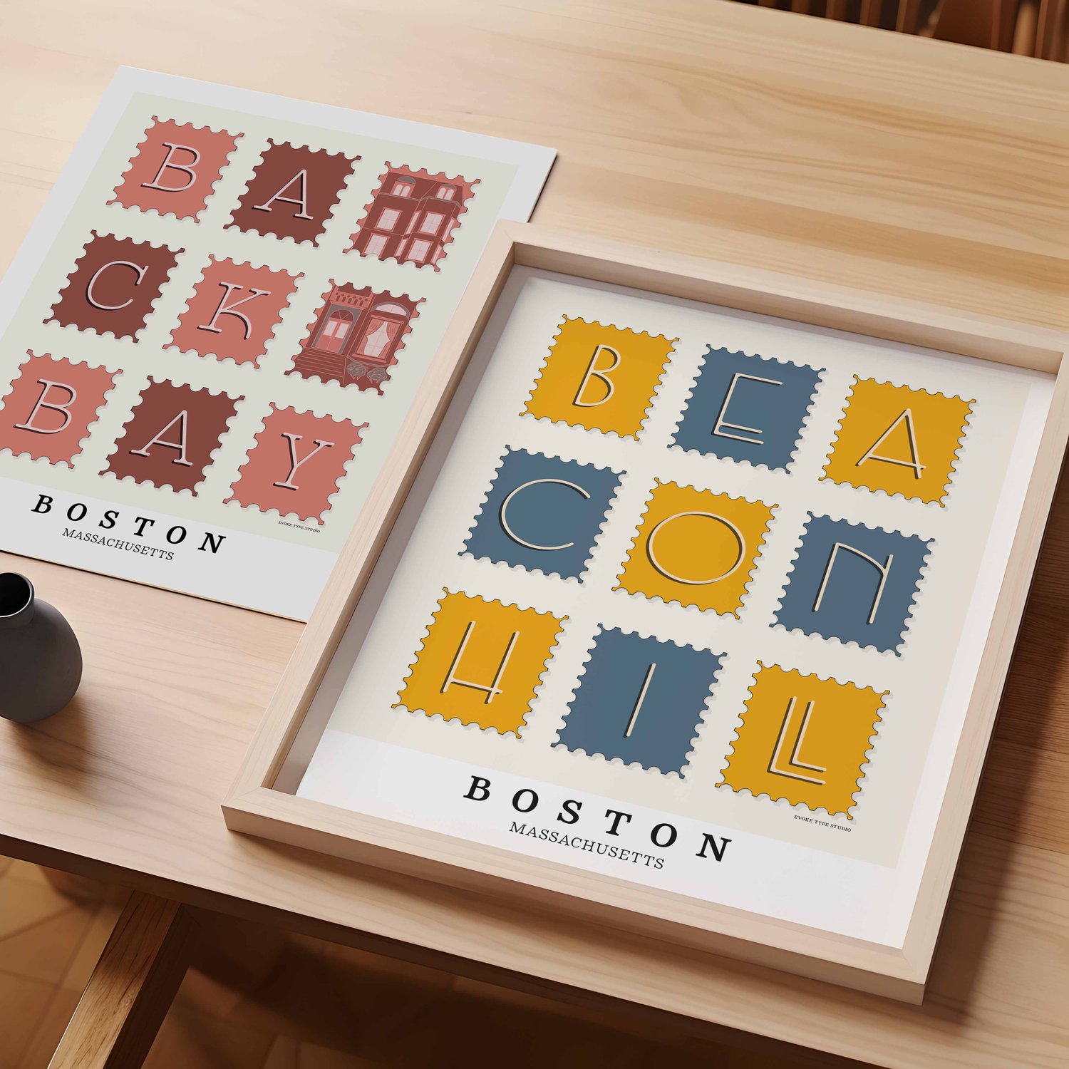 A table with an unframed artwork of Back Bay postage stamp art and a framed artwork of Beacon Hill postage stamp art