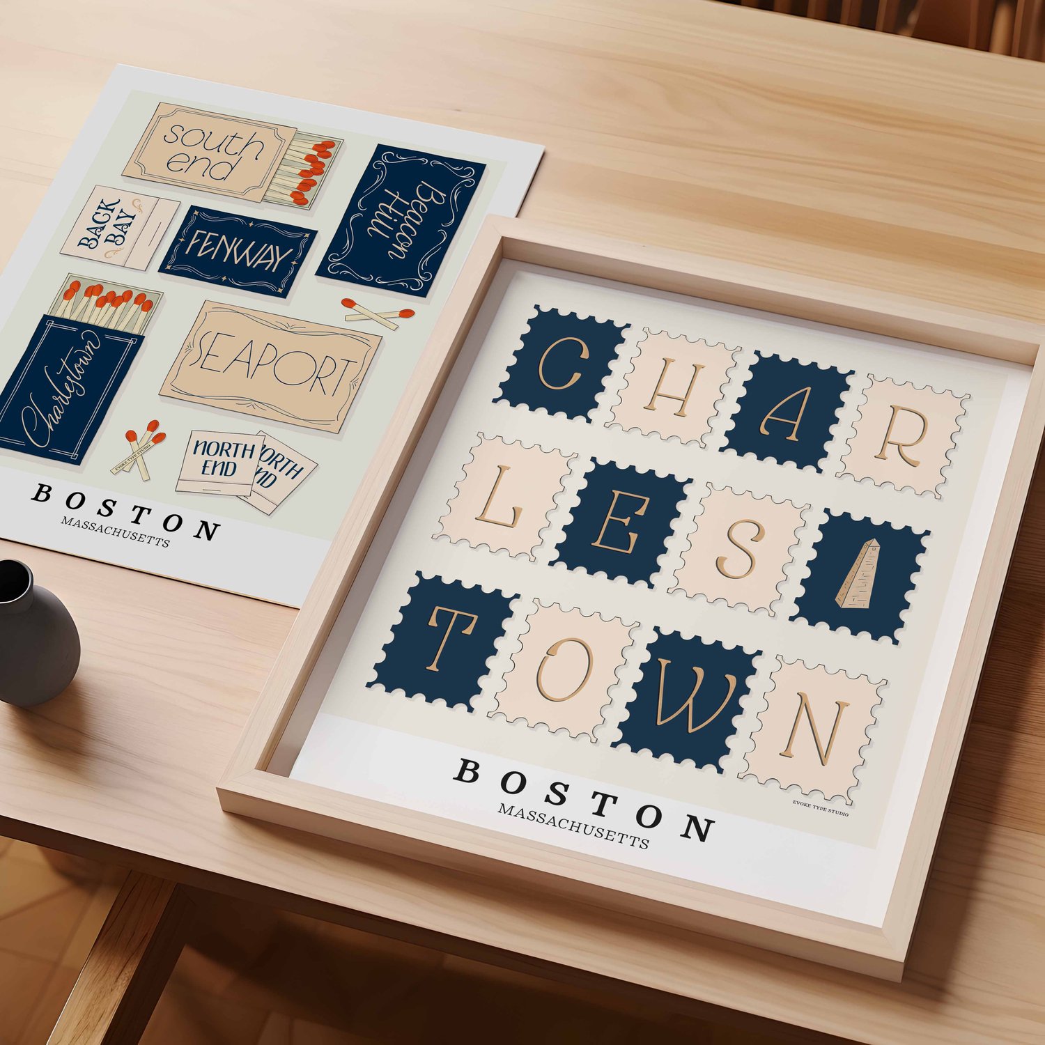 A table with an unframed artwork of Boston, Massachusetts matchbox art and a framed artwork of Charlestown postage stamp art