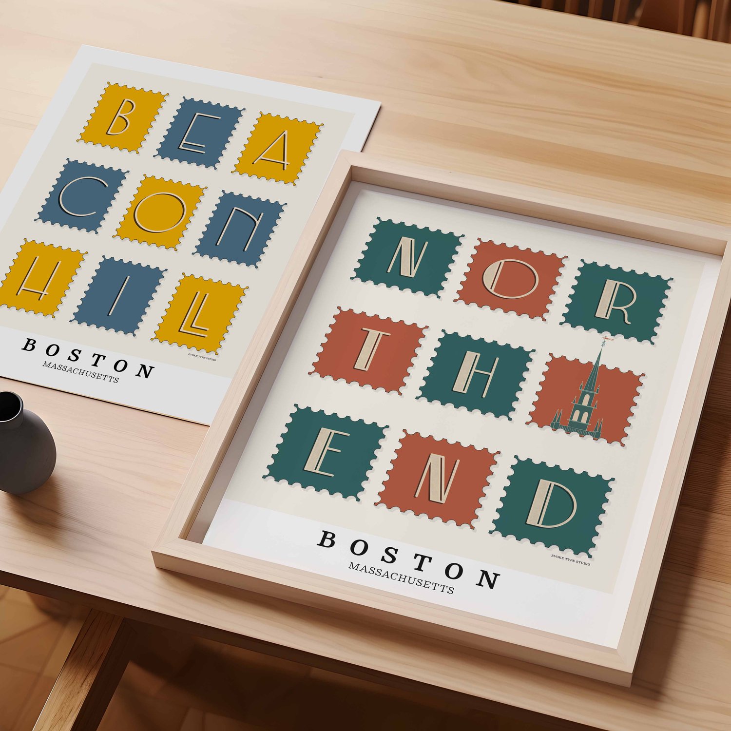 A table with an unframed artwork of Beacon Hill postage stamp art and a framed artwork of North End postage stamp art