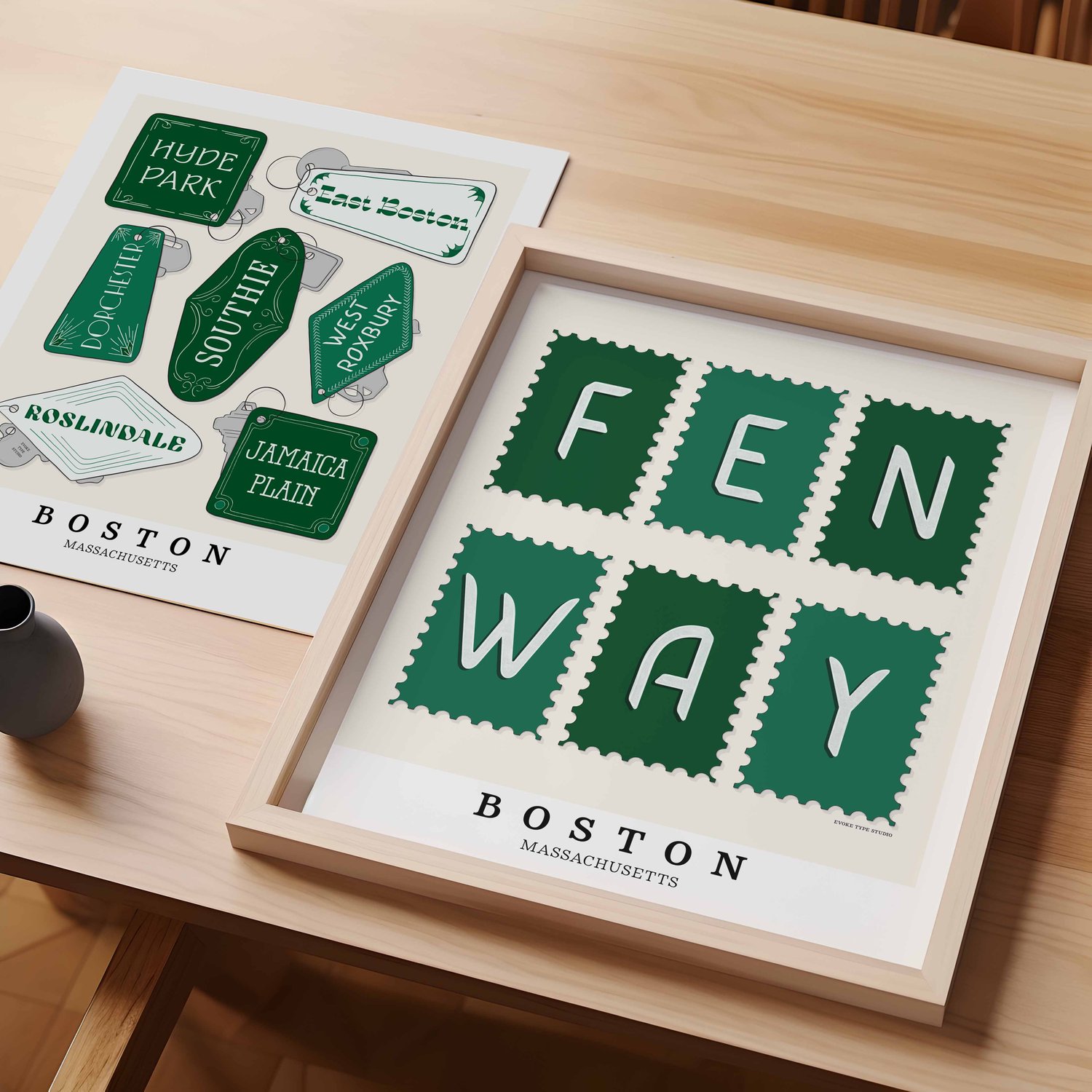 A table with an unframed artwork of Boston neighborhoods key fob art and a framed artwork of Fenway postage stamp art