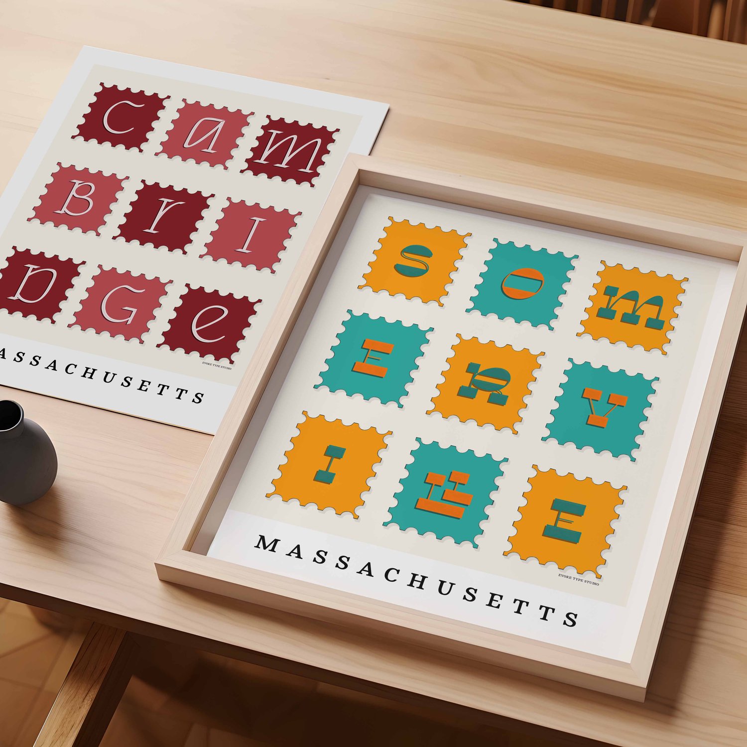 A table with an unframed artwork of Cambridge postage stamp art and a framed artwork of Somerville postage stamp art