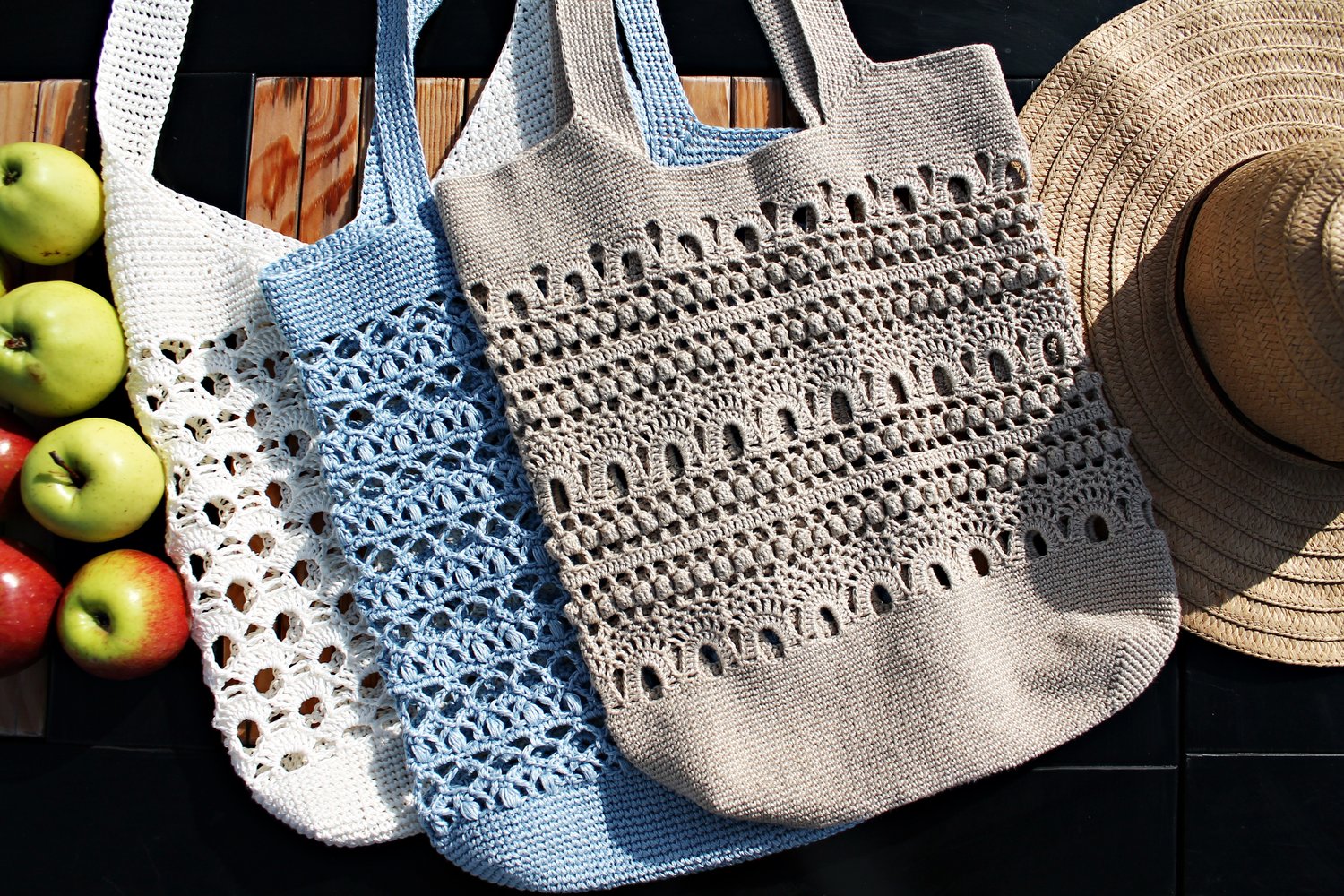 Three handmade crochet tote bags in beige, blue, and white, made with linen yarn and lace pattern. Designed by Kate Kosenko (Veselunka / ItWasYarn). Sunglasses and apples on the table nearby.