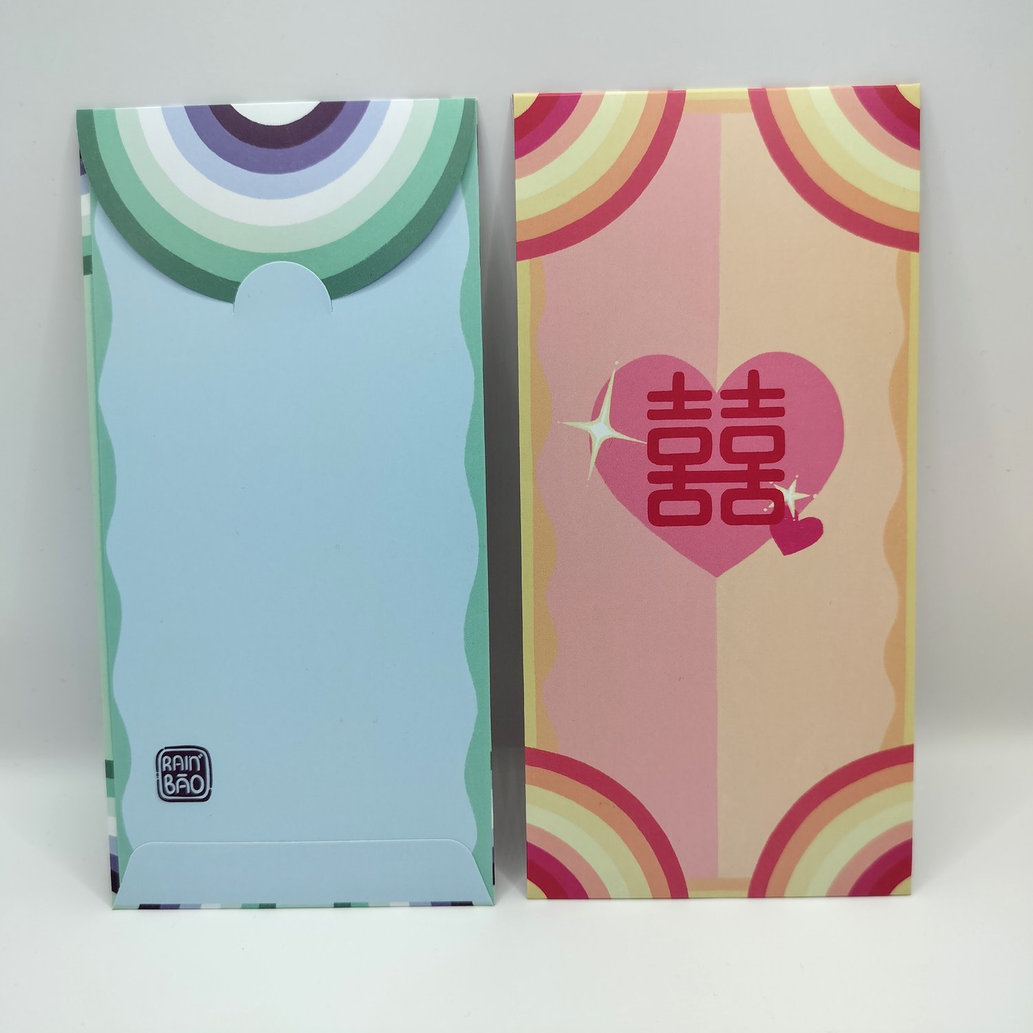Two money envelopes side by side - a blue envelope with green and blue rainbow arc design, next to a colorful envelope with pink and yellow rainbow stripes featuring a pink heart with Chinese double happiness characters.