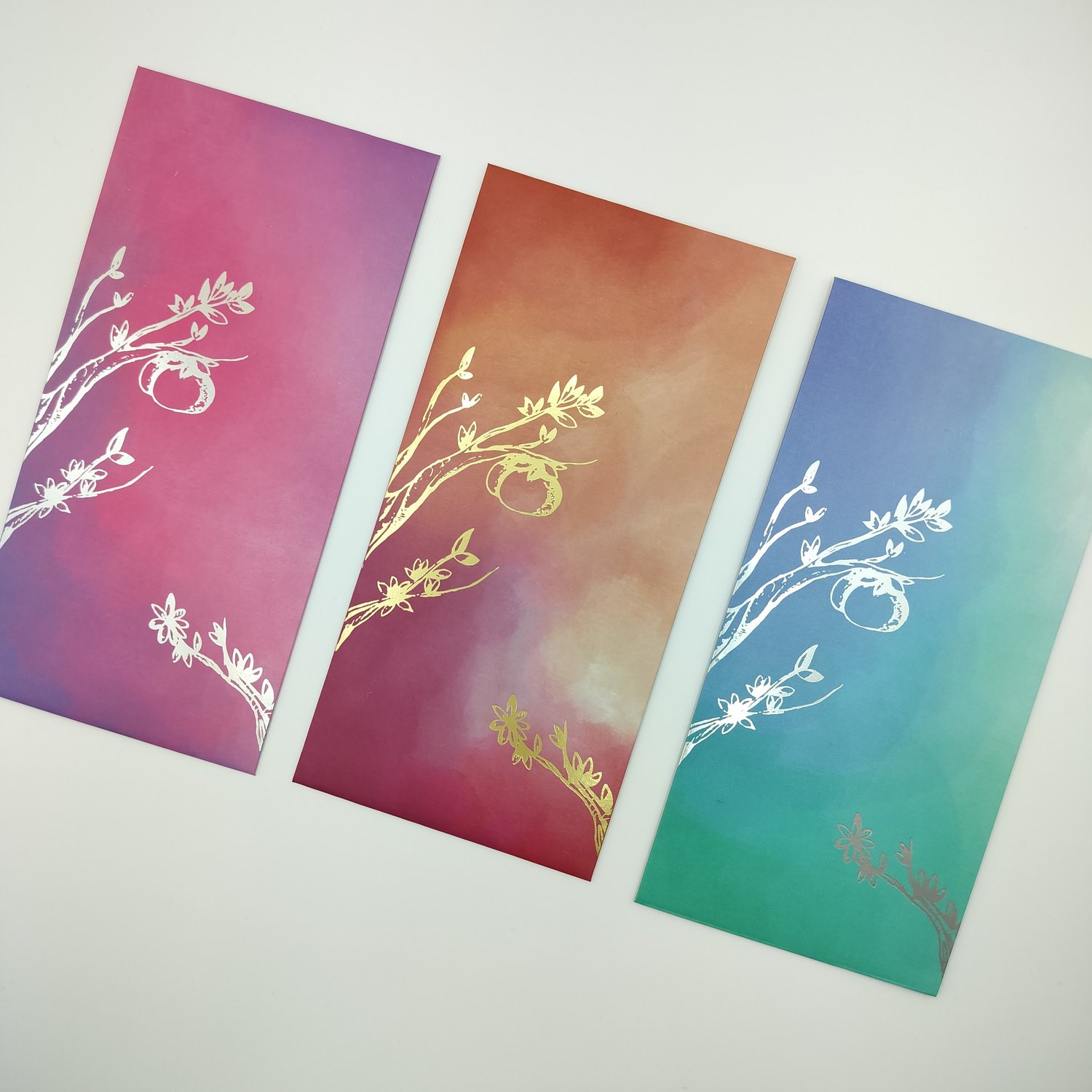 Three gradient envelopes with metallic foil botanical designs - pink with silver foil, orange with gold foil, and blue-green with silver foil.