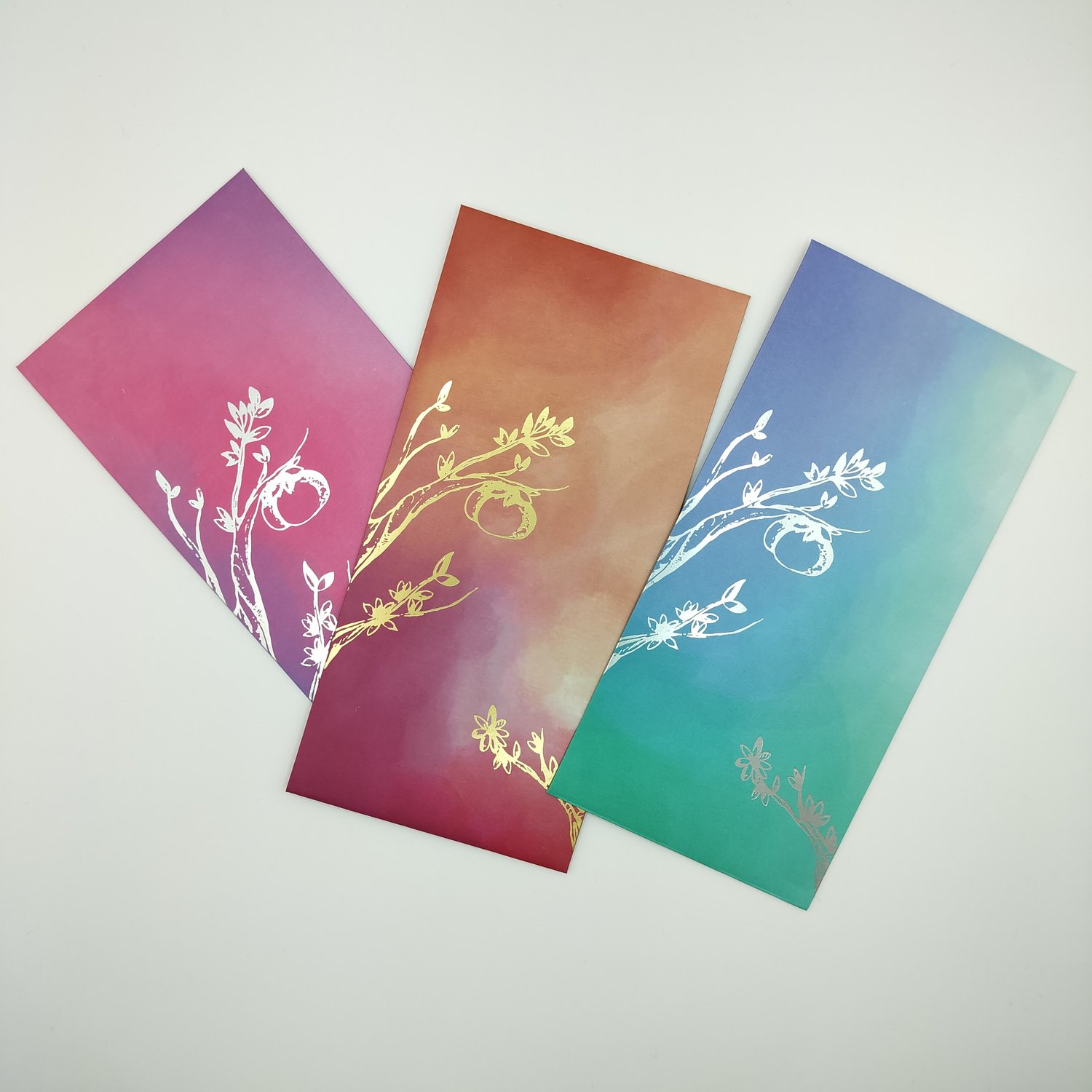 Three overlapping gradient envelopes with metallic foil botanical illustrations - a pink-to-purple card with silver foil branches and leaves, an orange-to-red version, and a blue-to-teal version.