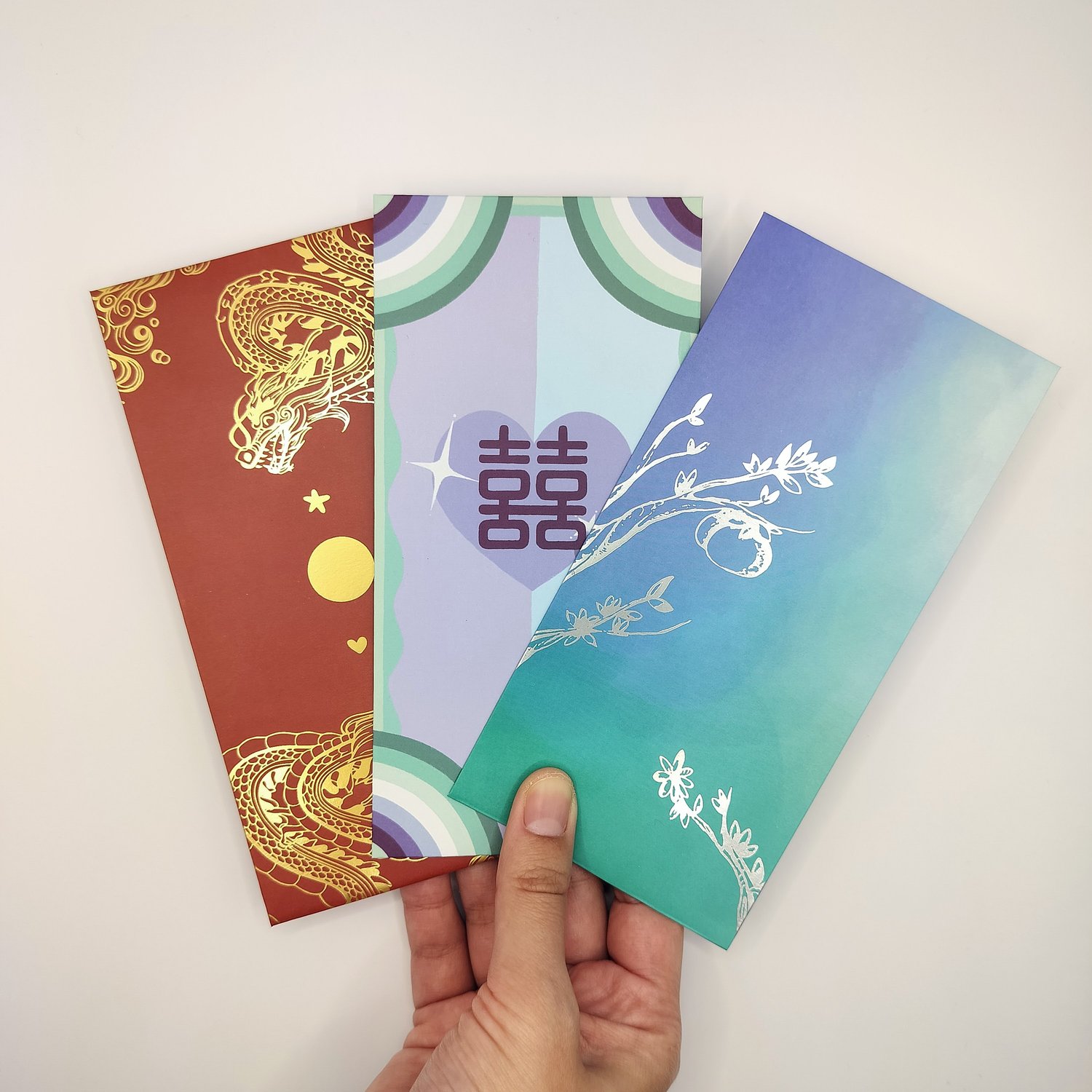 Hand holding three hongbaos- a red with gold dragon design, a pastel rainbow card with Chinese double happiness symbols, and a blue-to-teal gradient card with silver botanical foil designs.