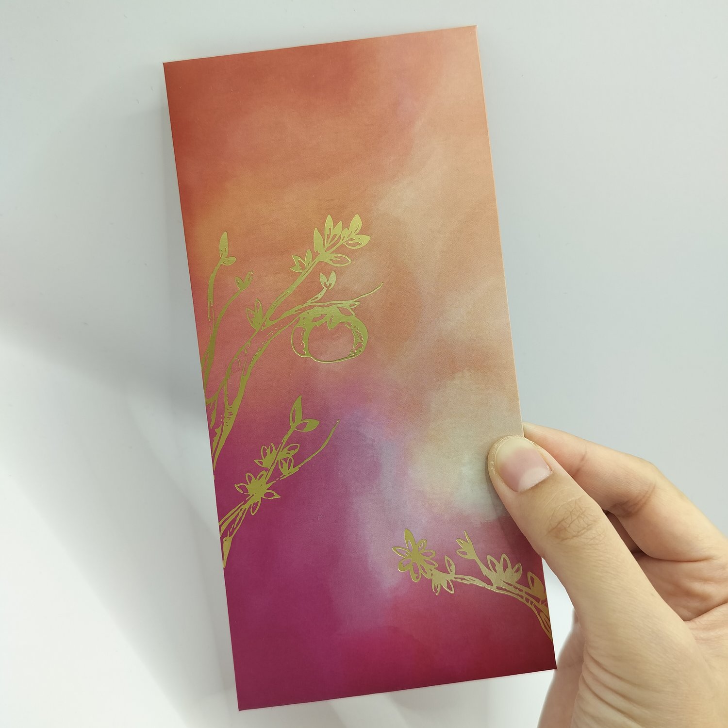 Hand holding a red, orange and pink gradient envelope with gold trim and small botanical motifs.