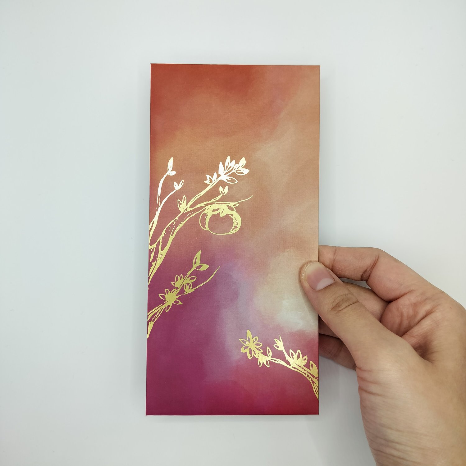 Hand holding a red, orange and pink gradient envelope with gold trim and small botanical motifs.
