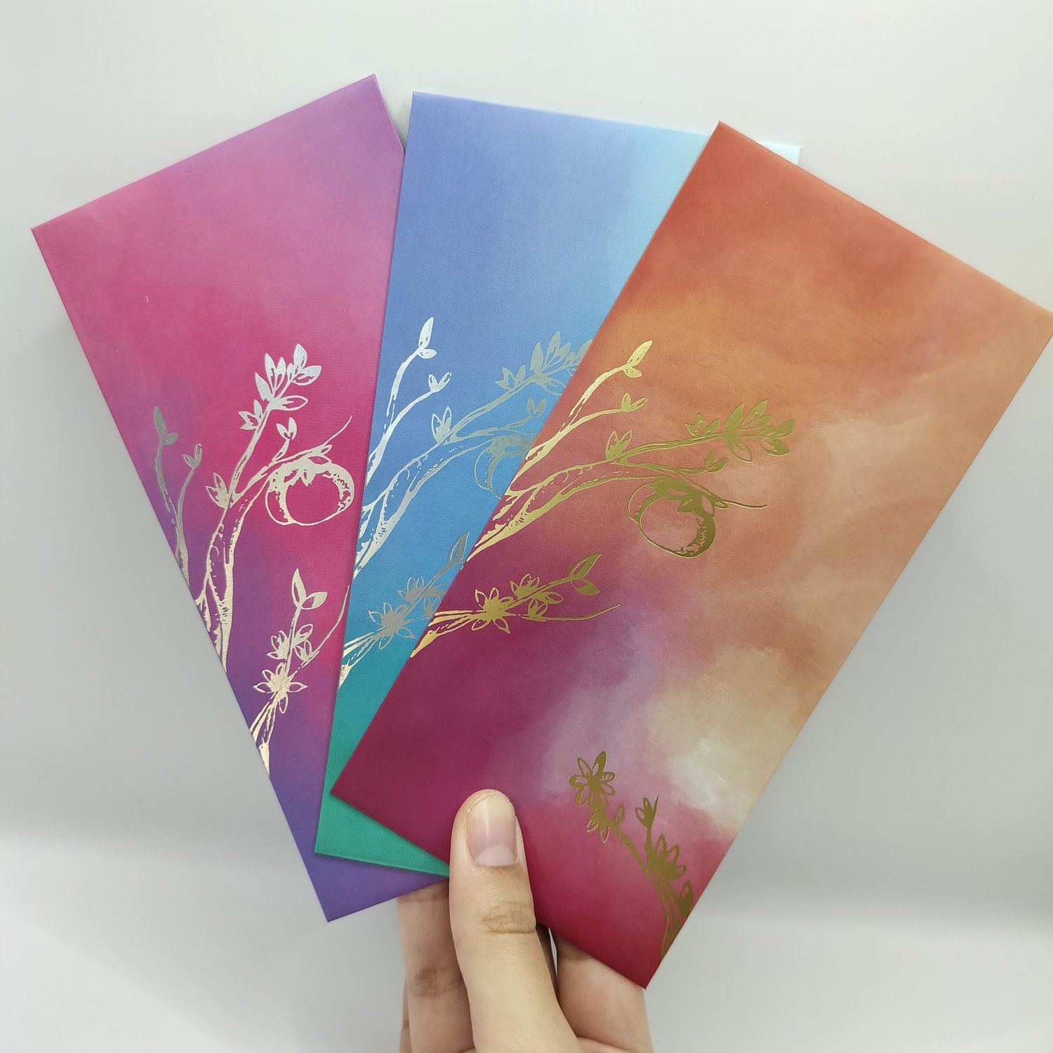 Hand holding three fanned gradient envelopes with metallic foil botanical designs - pink-to-purple with silver foil, blue-to-teal with silver foil, and orange-to-red with gold foil branch motifs.