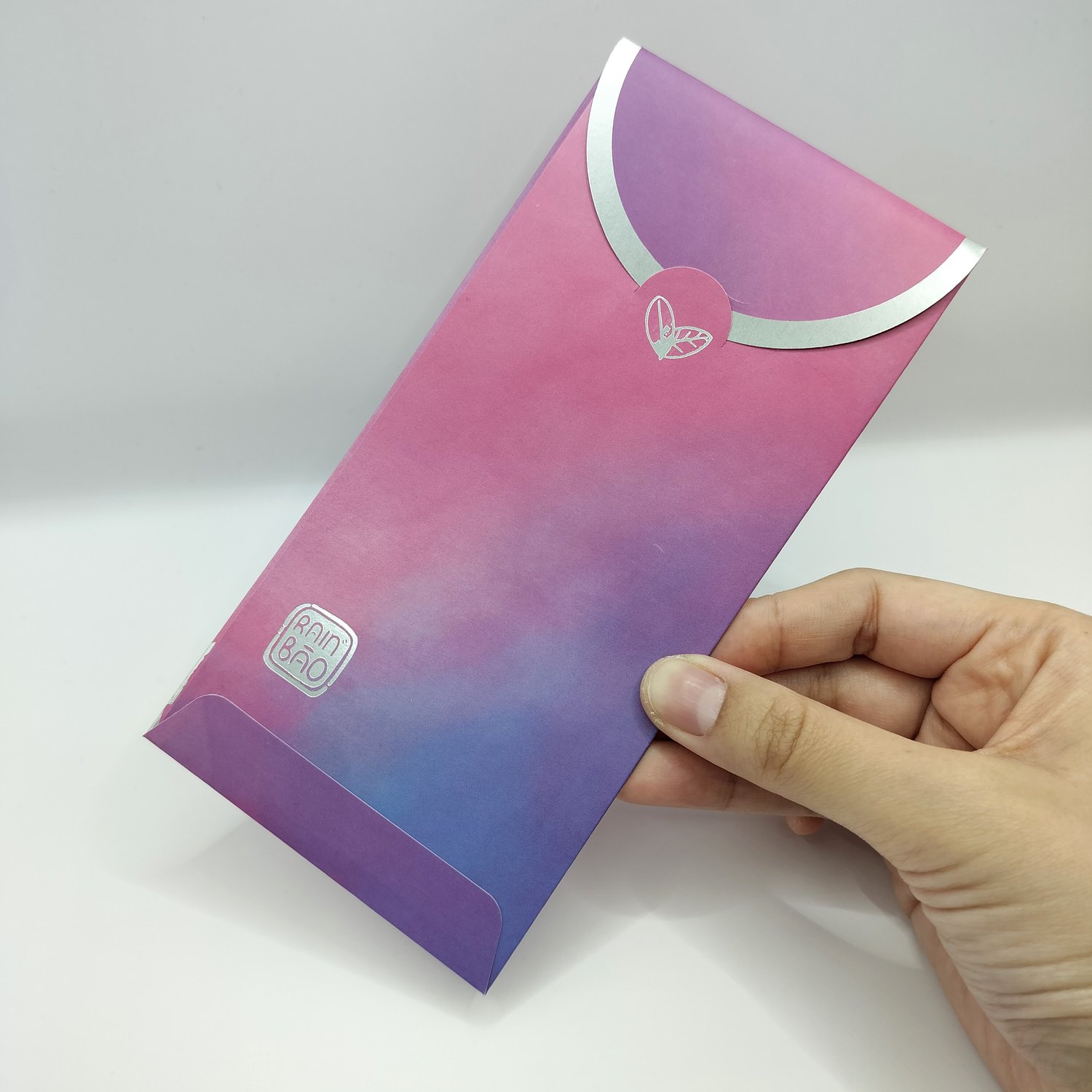 Hand holding a pink-to-purple gradient envelope with silver curved design elements and small botanical motifs, featuring a logo in the bottom corner.