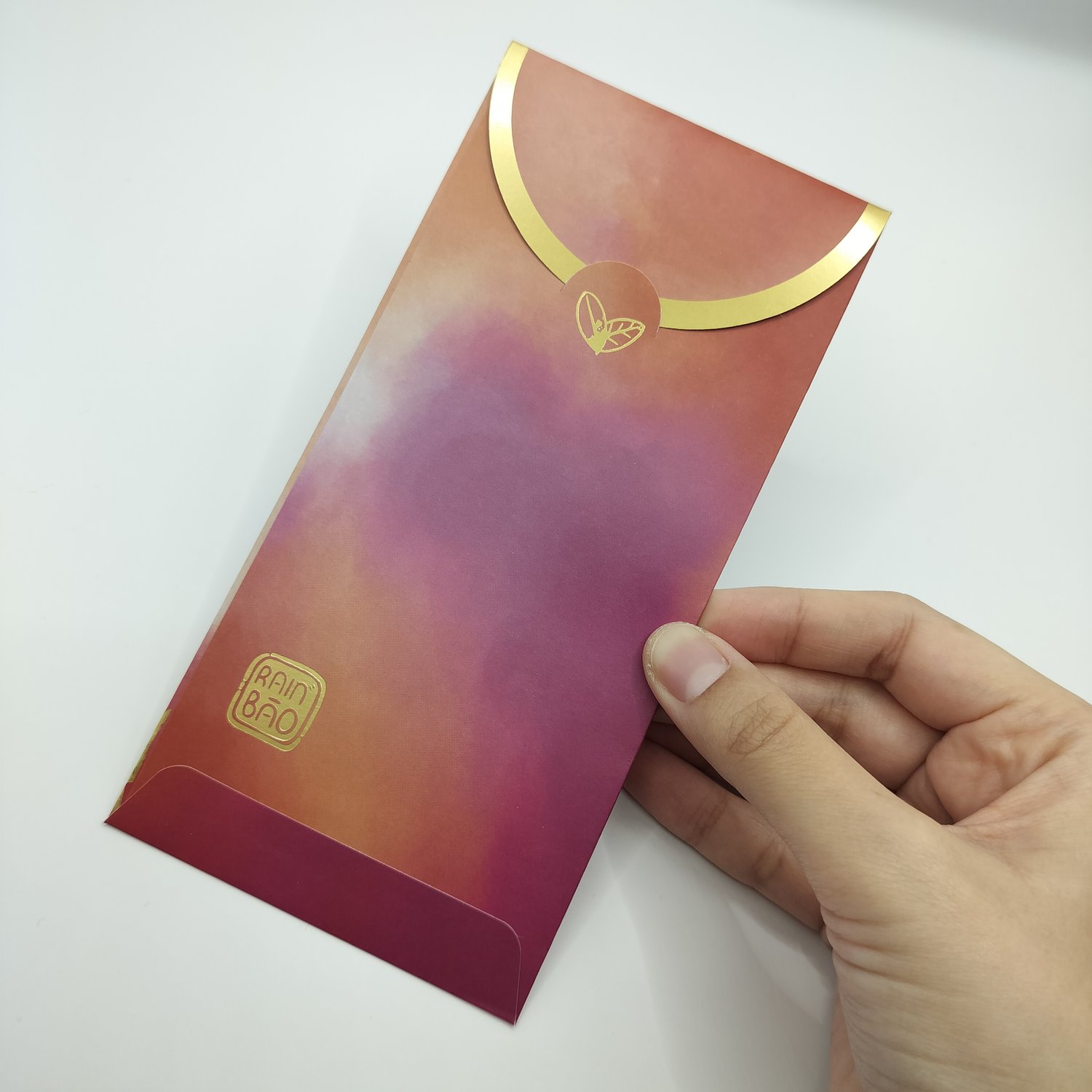 Hand holding a red-to-orange gradient envelope with gold curved design elements and small botanical motifs, featuring a logo in the bottom corner.