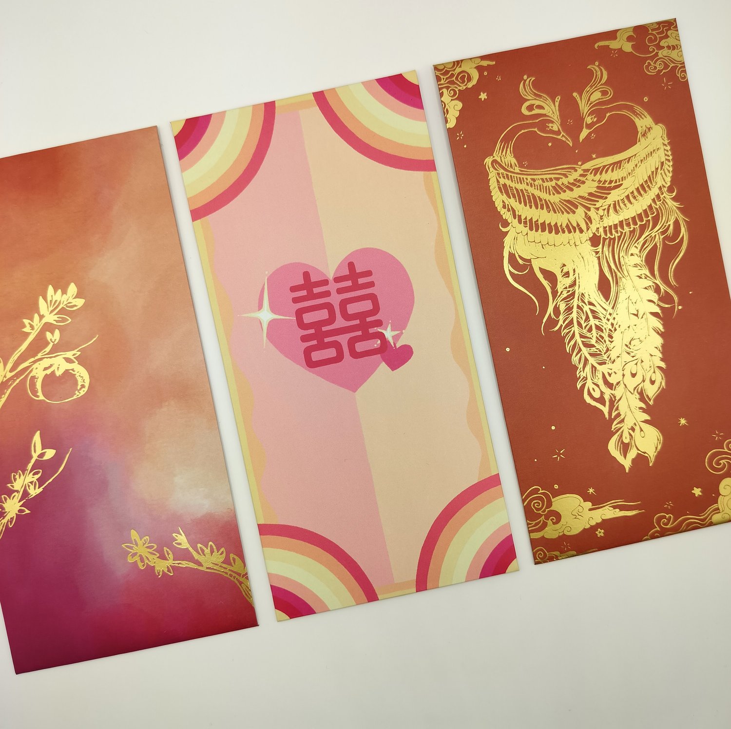 Three hongbao arranged in a row - an orange-to-red gradient envelope with gold botanical foil design, a pink and yellow rainbow striped envelope with double happiness heart, and a red envelope with elaborate gold phoenix design.
