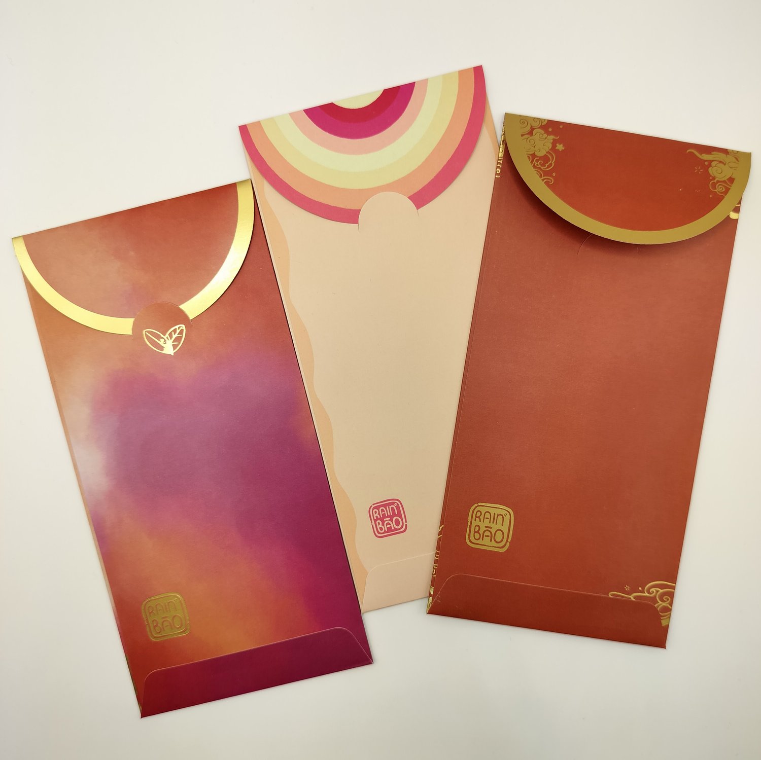 The backs of three hongbao arranged in a row - an orange-to-red gradient envelope with gold foil trim, a pink and yellow rainbow striped envelope and a red envelope with gold foil trim and small cloud details.