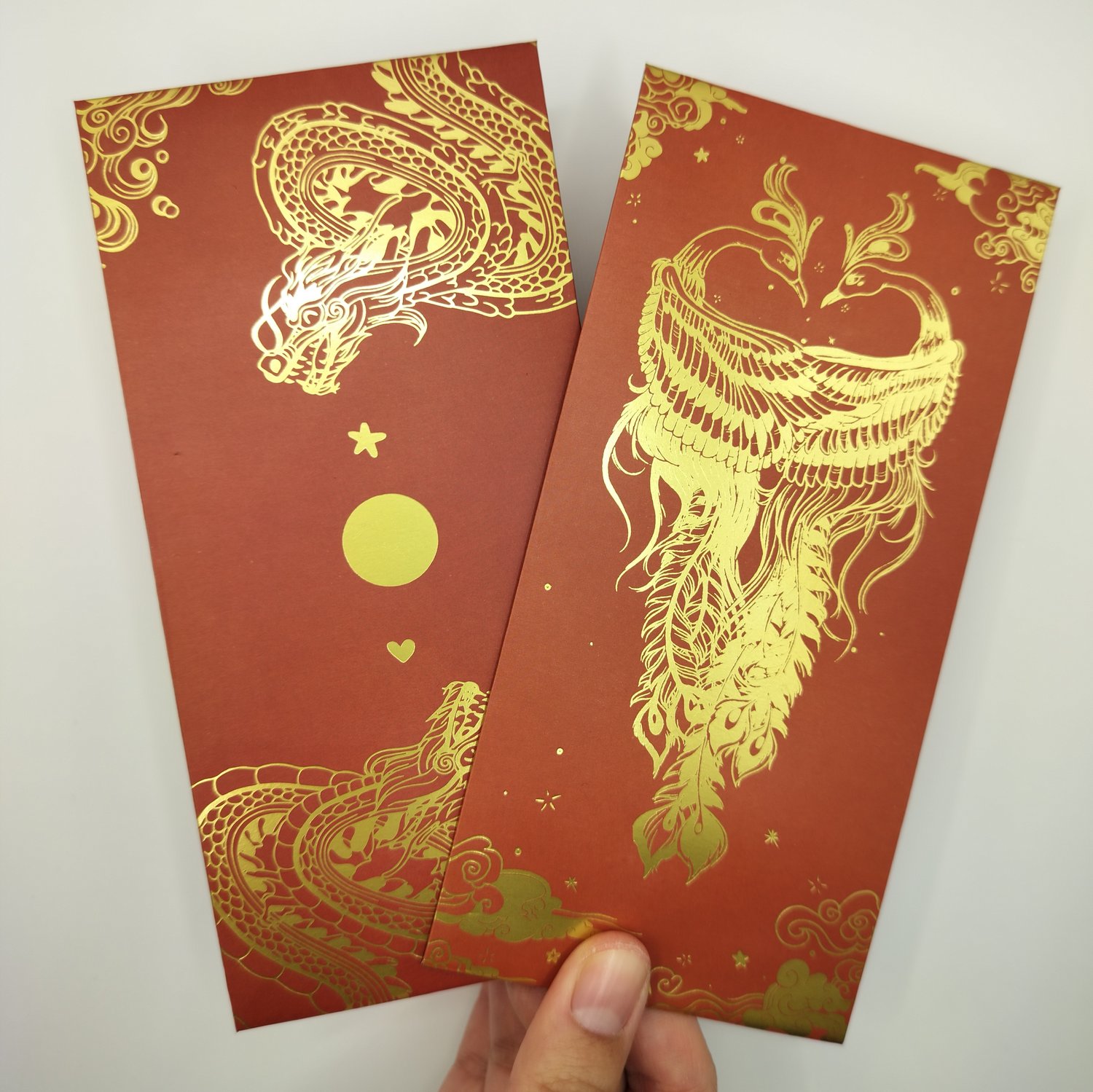 Hand holding two red hongbaos with gold foil designs - one featuring dragons with decorative elements like stars and circles, and another with an two phoenixes surrounded by clouds and ornamental details.