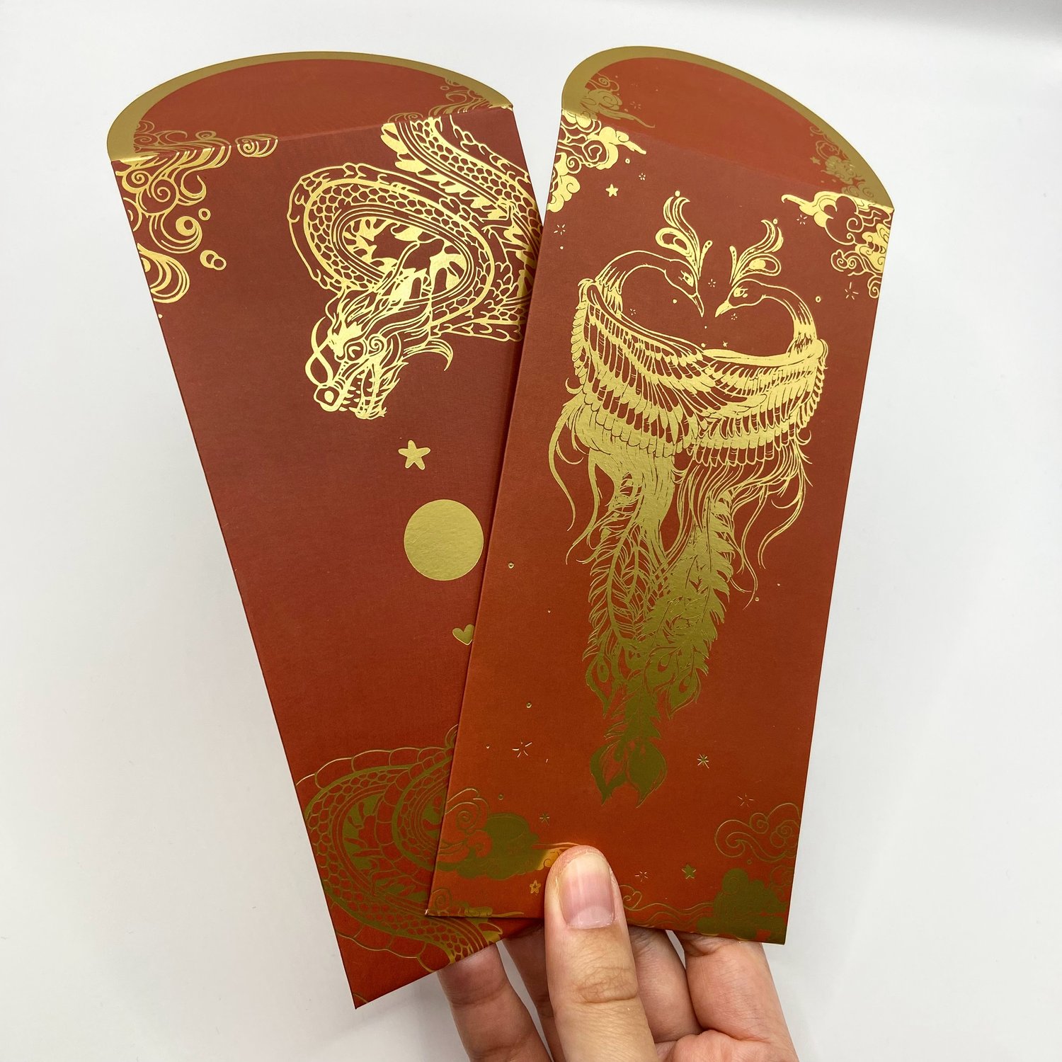 Hand holding two red hongbaos with gold foil designs - one featuring dragons with decorative elements like stars and circles, and another with an two phoenixes surrounded by clouds and ornamental details.
