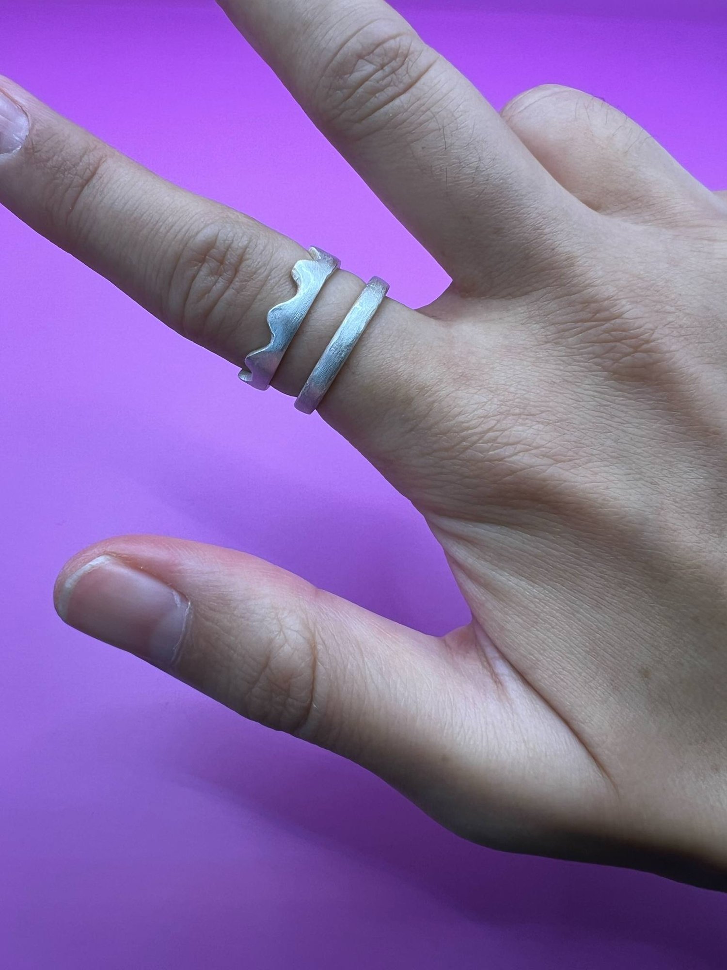 A hand wearing two silver rings. One has a wavy, scalloped edge and the other is a plain band.