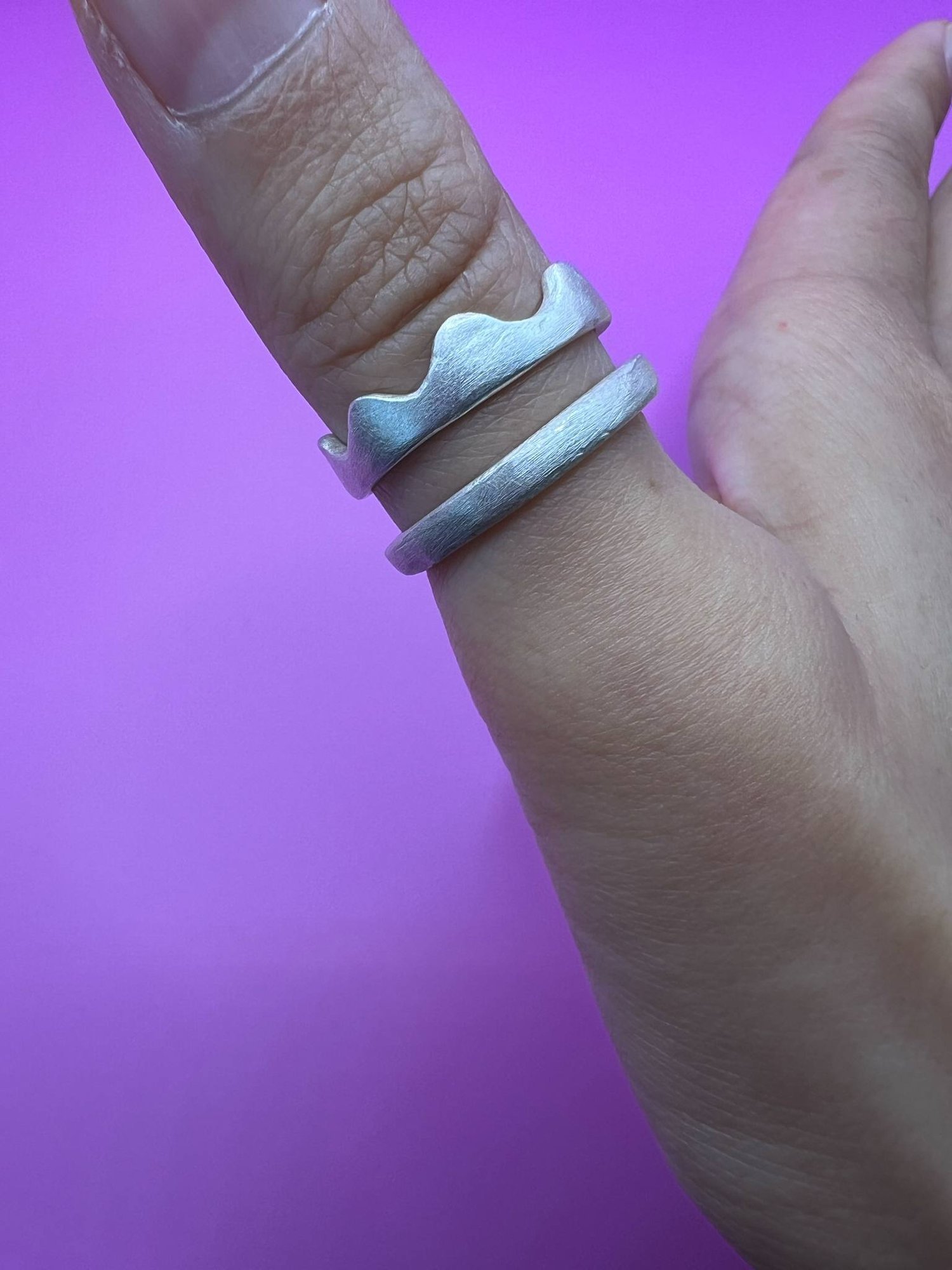 A thumb wearing two stacked silver rings. One has a wavy, scalloped edge and the other is a plain band.
