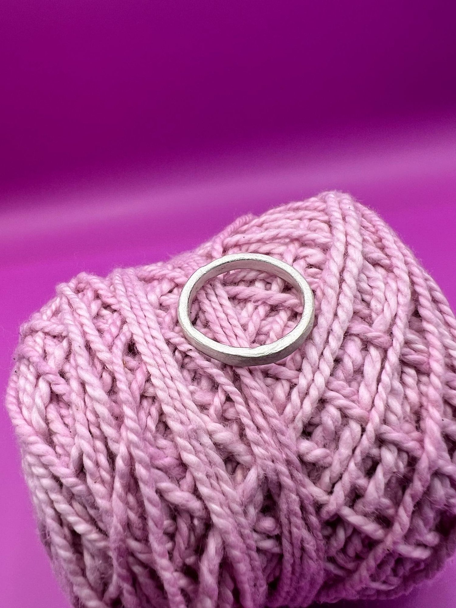 Side profile of a simple silver band ring nestled in pink yarn against a vibrant magenta background.