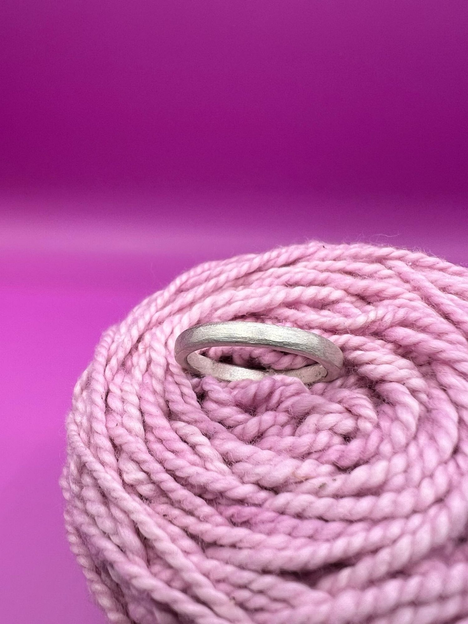 A simple silver band ring nestled in pink yarn against a vibrant magenta background.
