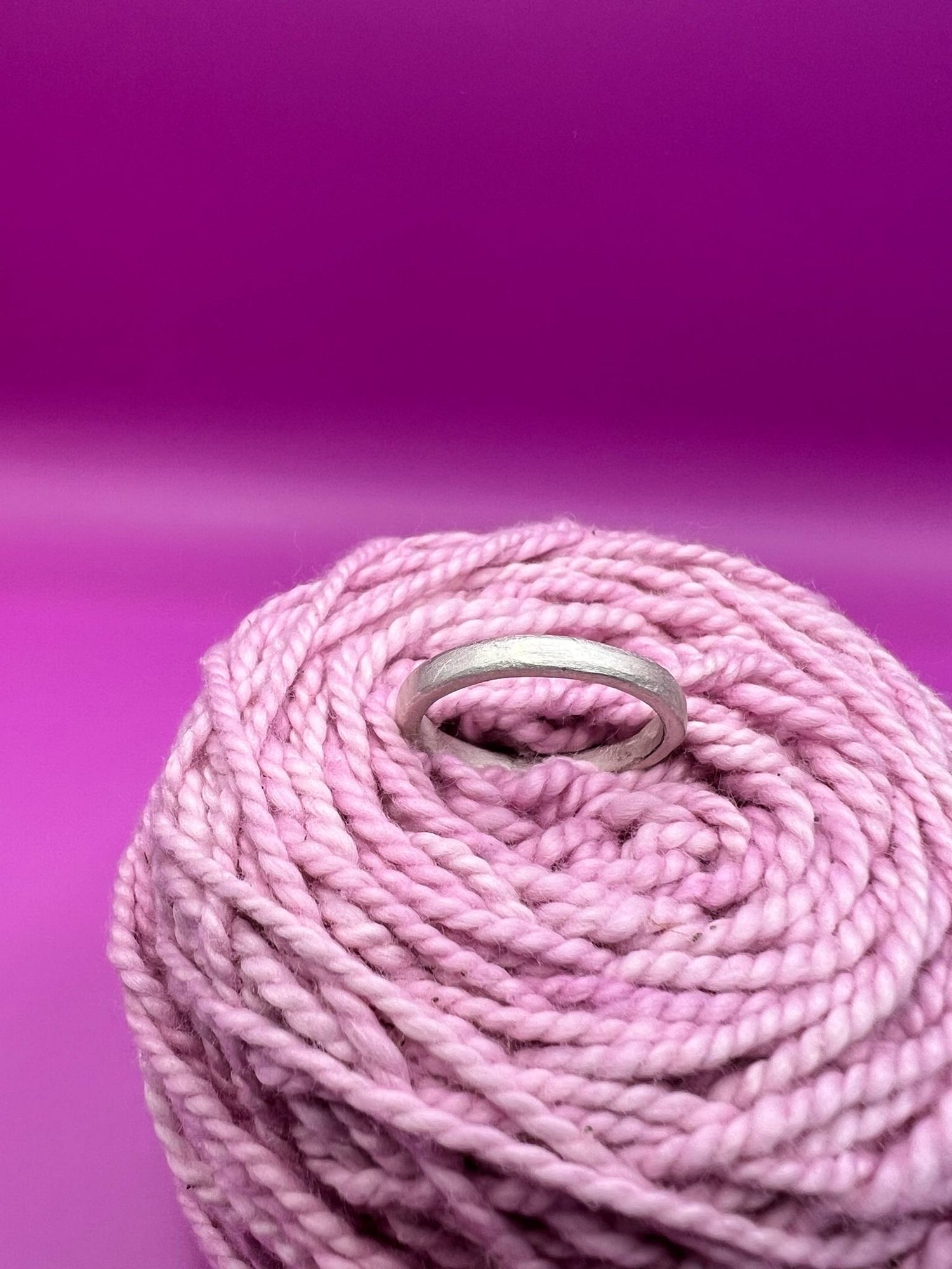 A simple silver band ring nestled in pink yarn against a vibrant magenta background.