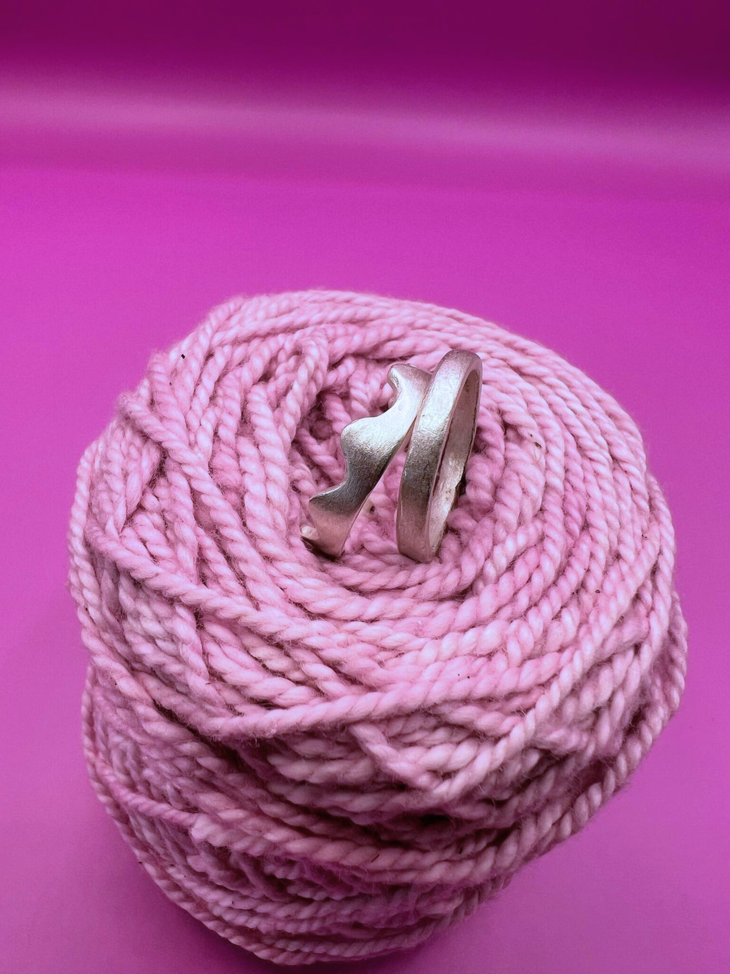 Two silver rings nestled in coiled pink yarn against a vibrant magenta background. One has a wavy, scalloped edge and the other is a plain band.