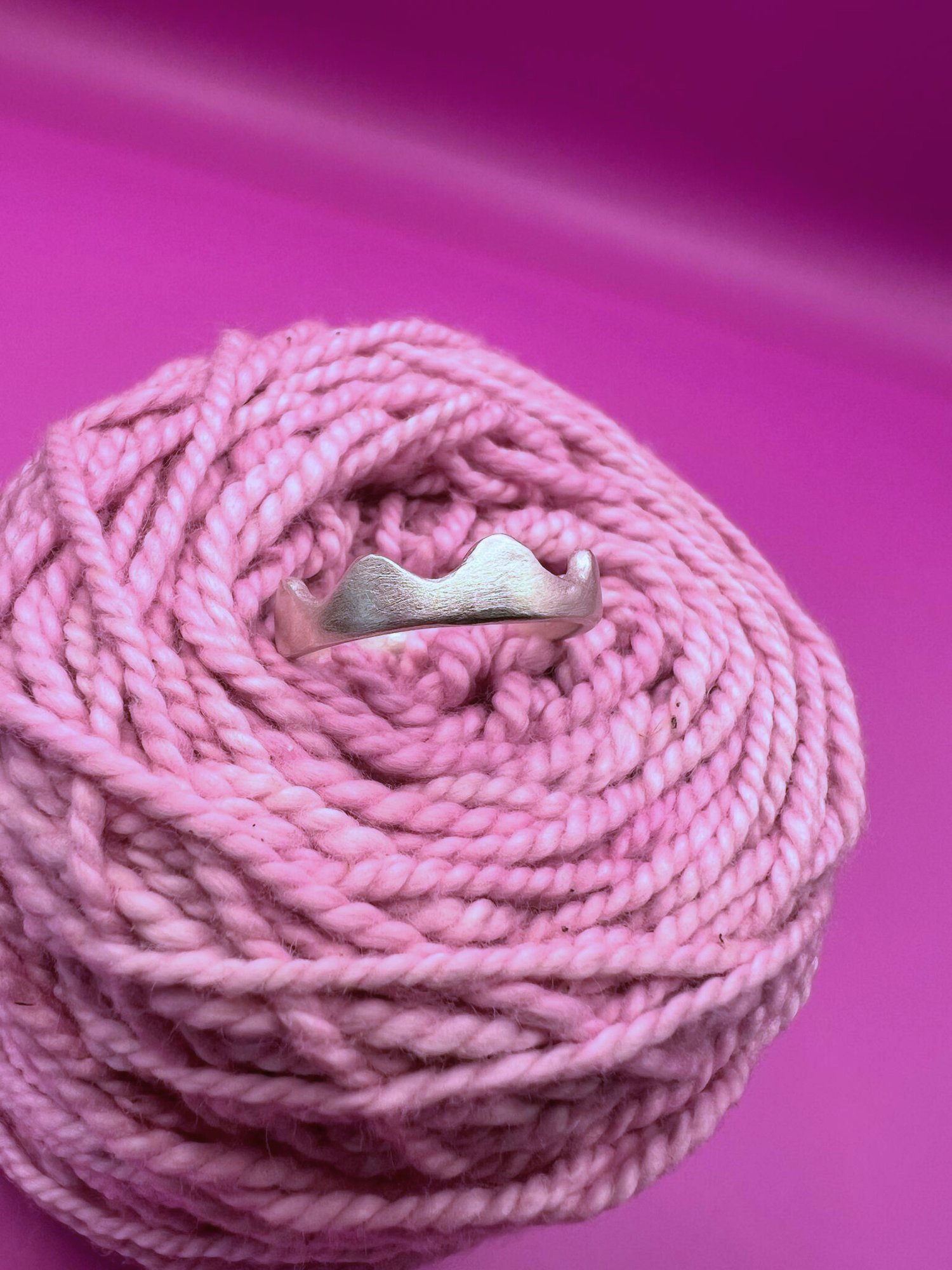 Side profile of a silver ring with wavy, scalloped edges nestled in coiled pink yarn against a vibrant magenta background.