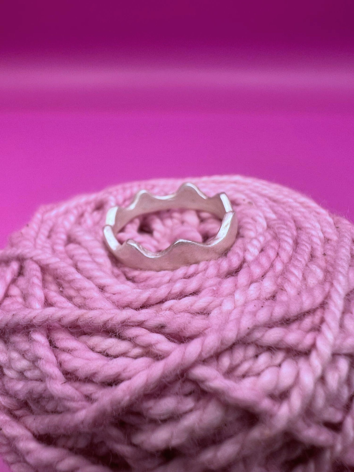Silver ring with wavy, scalloped edges nestled in coiled pink yarn against a vibrant magenta background.