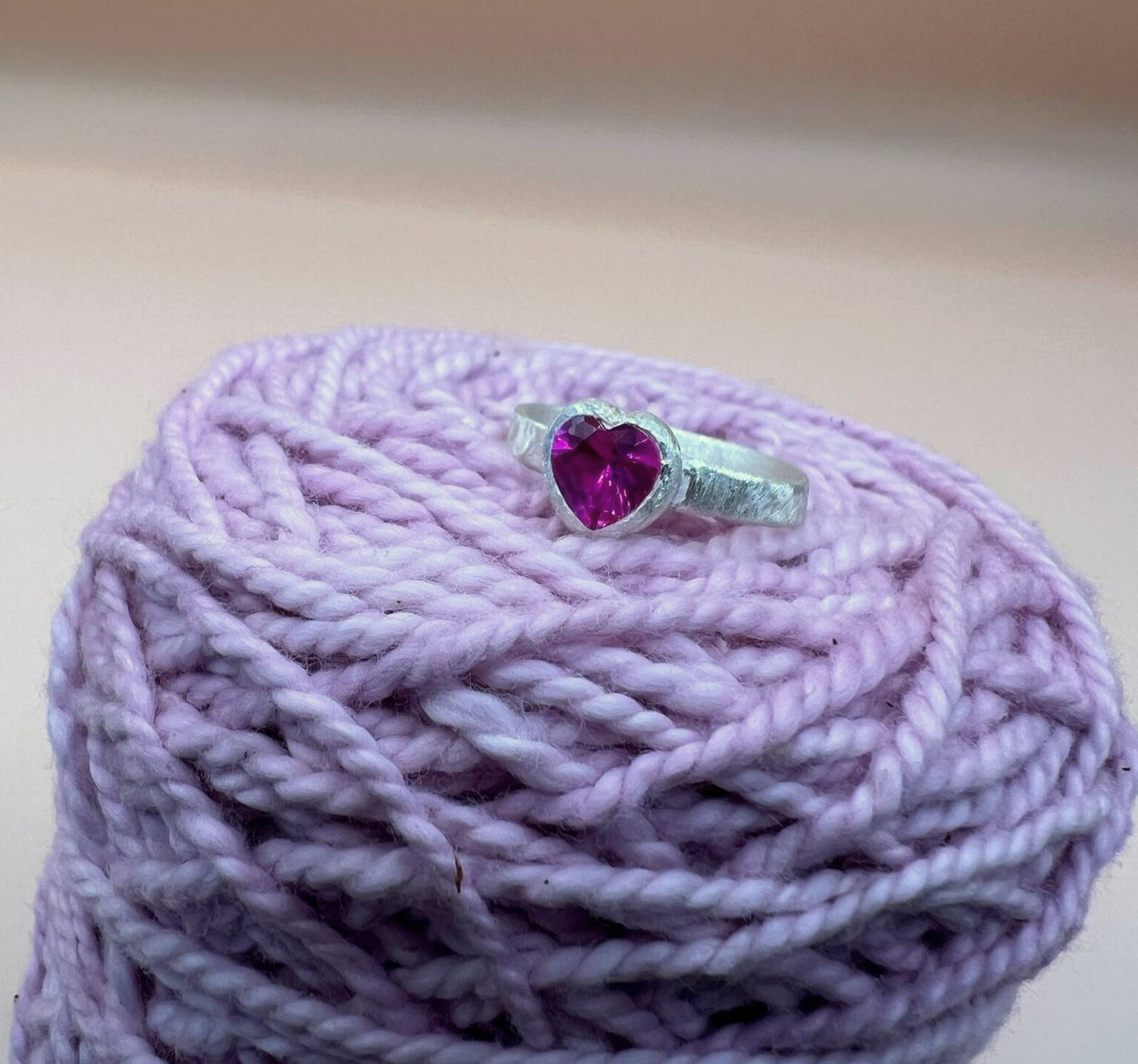 Silver ring with a textured band featuring a heart-shaped pink ruby in a bezel setting sitting on some pink yarn.