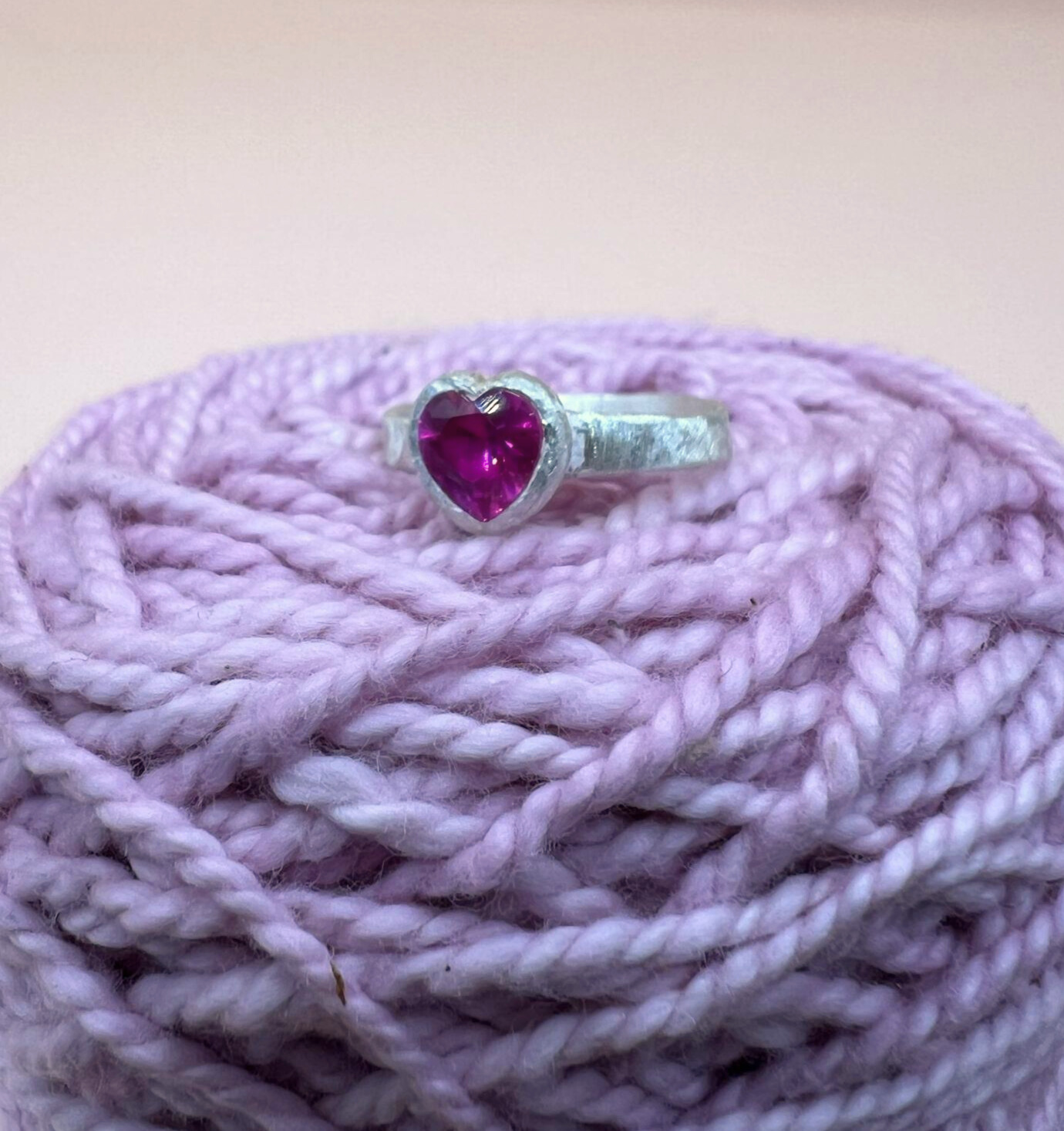 Silver ring with a textured band featuring a heart-shaped pink ruby in a bezel setting sitting on some pink yarn.