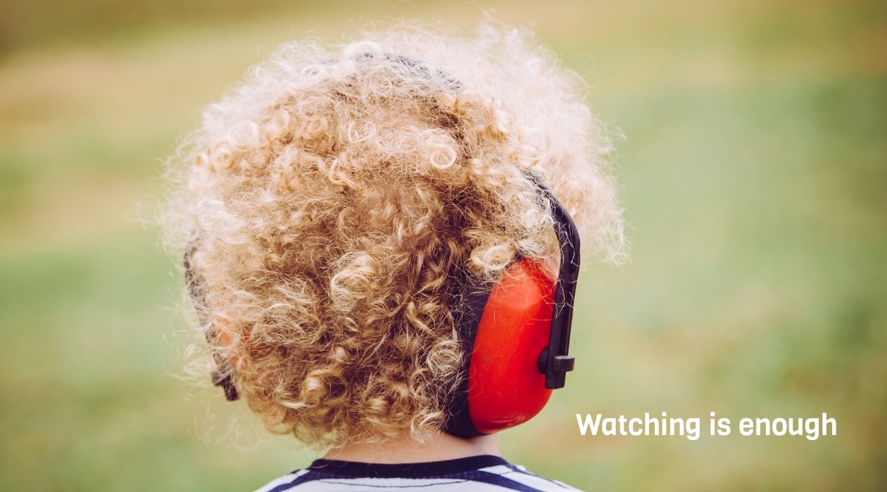 A young child with curly blonde hair is shown from behind, wearing large red ear defenders. They are standing outdoors on grass, with a soft green and yellow blurred background. The child appears to be observing quietly. In the bottom right corner, white 
