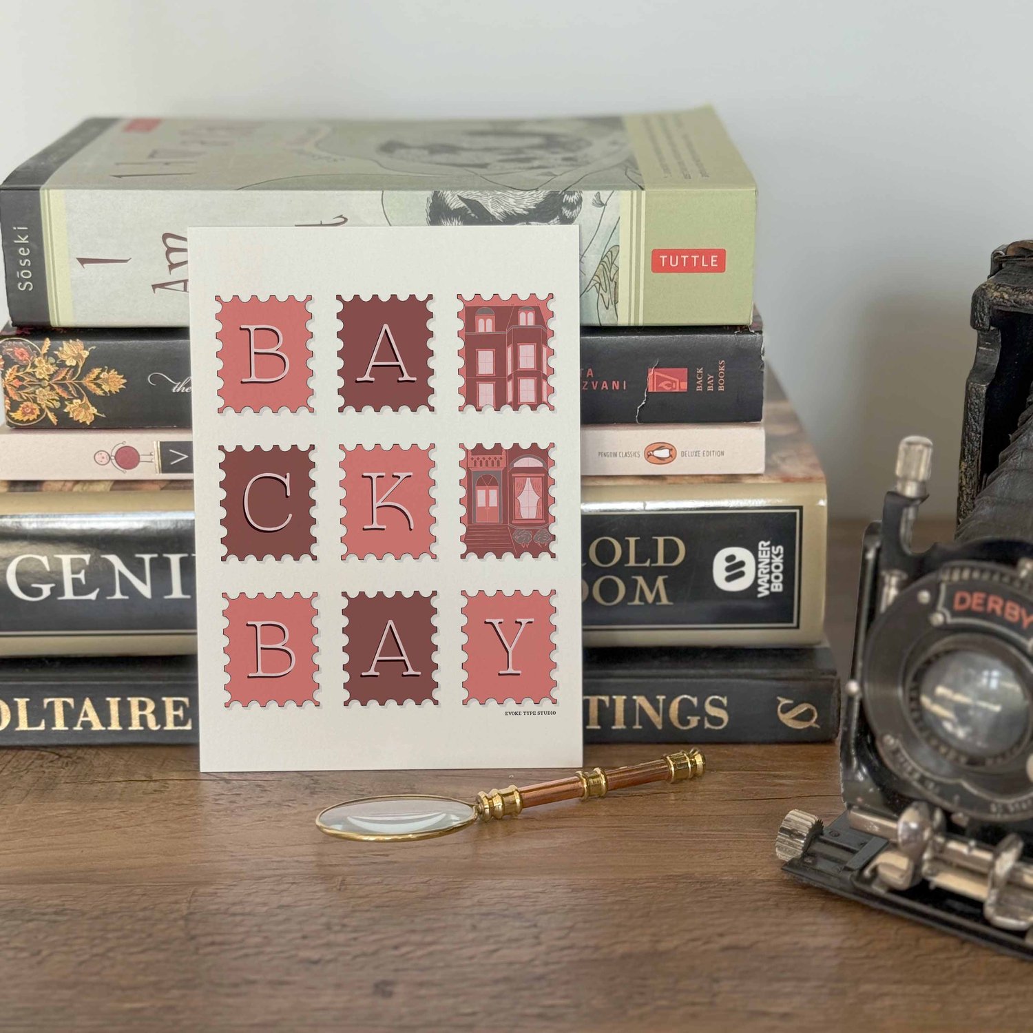 A notecard placed in front of a stack of books with a vintage camera and a small magnifying glass placed nearby. The artwork on the notecard is of postage stamps featuring the Back Bay neighborhood in Boston, Massachusetts drawn in a unique lettering styl