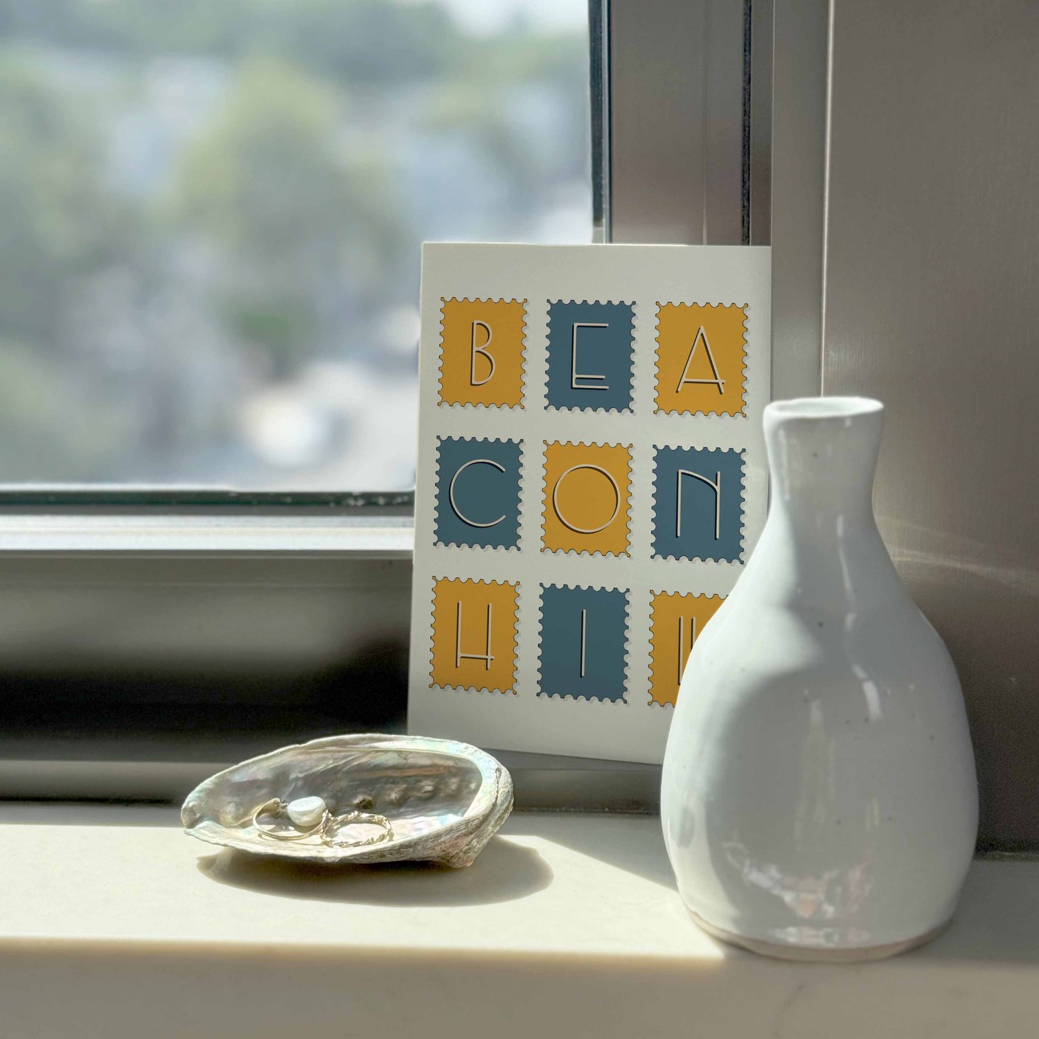 A notecard sitting on a windowsill surrounding by a white vase and a pretty shell with jewelry placed inside.