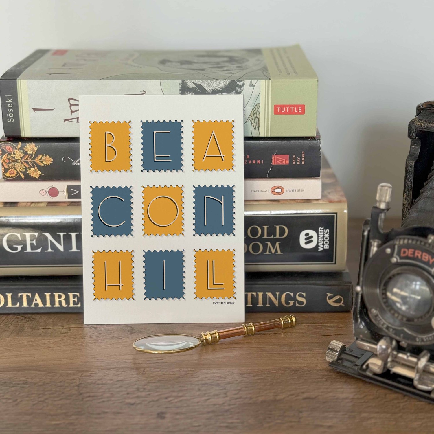 A notecard placed in front of a stack of books with a vintage camera and a small magnifying glass placed nearby. The artwork on the notecard is of postage stamps featuring the Beacon Hill neighborhood in Boston, Massachusetts drawn in a unique lettering s