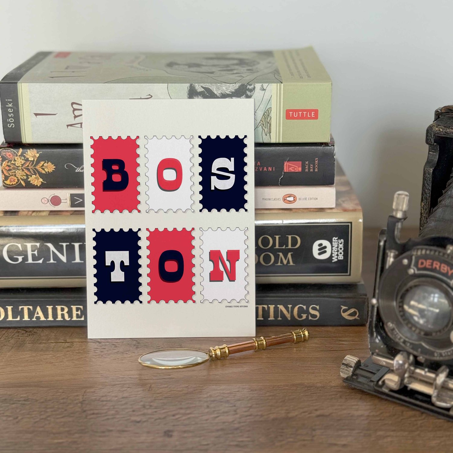 A notecard placed in front of a stack of books with a vintage camera and a small magnifying glass placed nearby. The artwork on the notecard is of postage stamps featuring Boston, Massachusetts drawn in a unique lettering style. Artwork has a red, white a