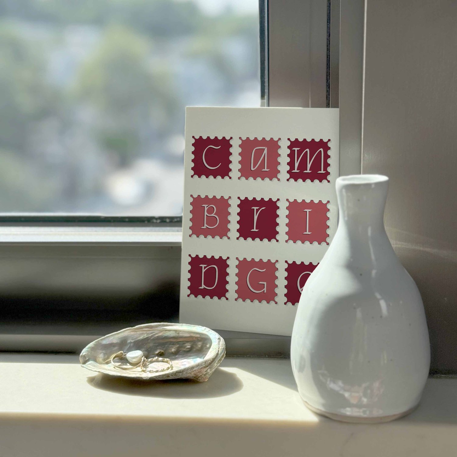 A notecard sitting on a windowsill surrounding by a white vase and a pretty shell with jewelry placed inside.