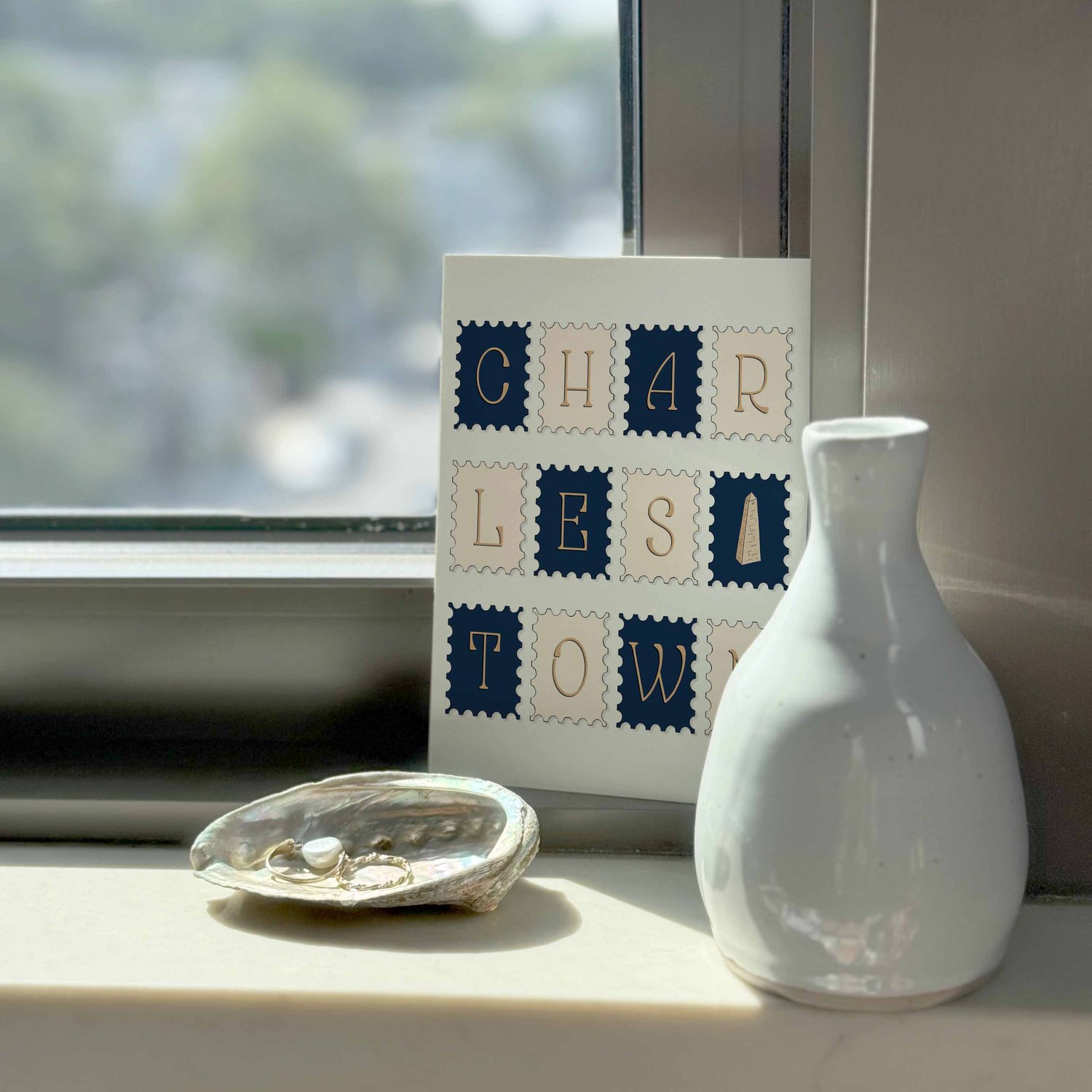 A notecard sitting on a windowsill surrounding by a white vase and a pretty shell with jewelry placed inside.