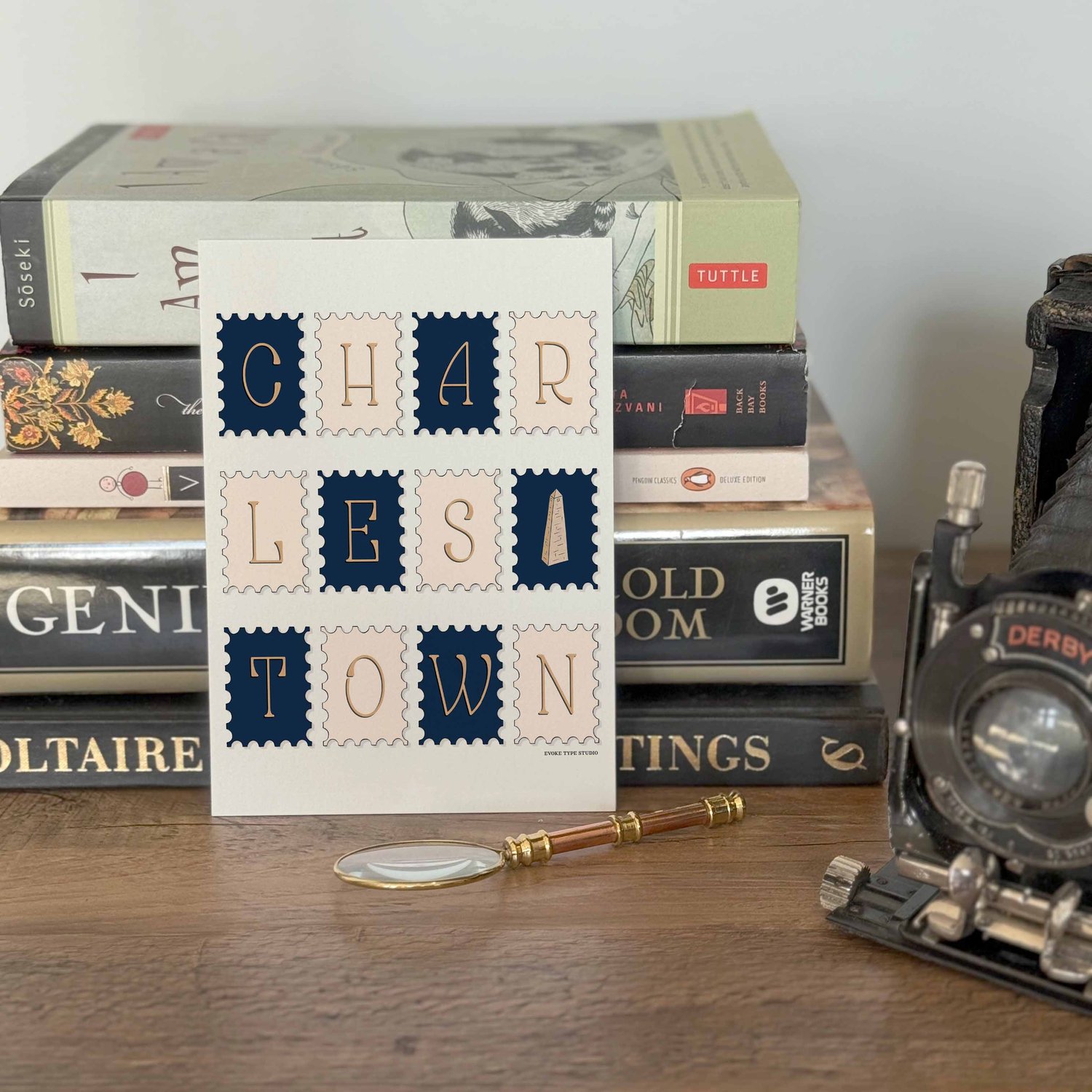 A notecard placed in front of a stack of books with a vintage camera and a small magnifying glass placed nearby. The artwork on the notecard is of postage stamps featuring the Charlestown neighborhood in Boston, Massachusetts drawn in a unique lettering s