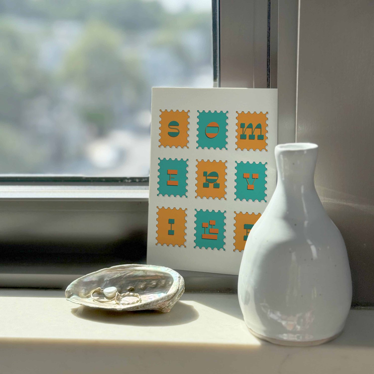 A notecard sitting on a windowsill surrounding by a white vase and a pretty shell with jewelry placed inside.