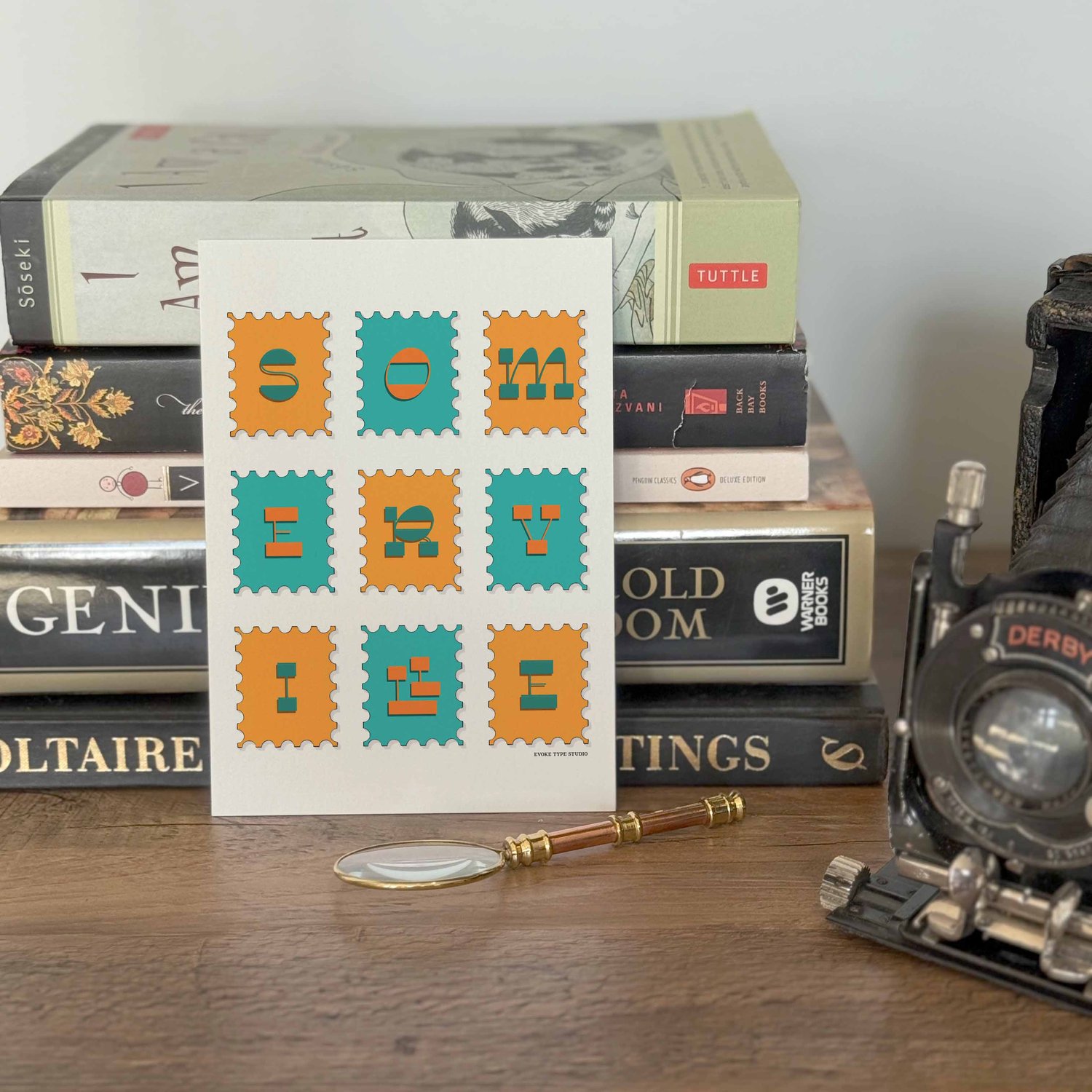 A notecard placed in front of a stack of books with a vintage camera and a small magnifying glass placed nearby. The artwork on the notecard is of postage stamps featuring Somerville, Massachusetts drawn in a unique lettering style.