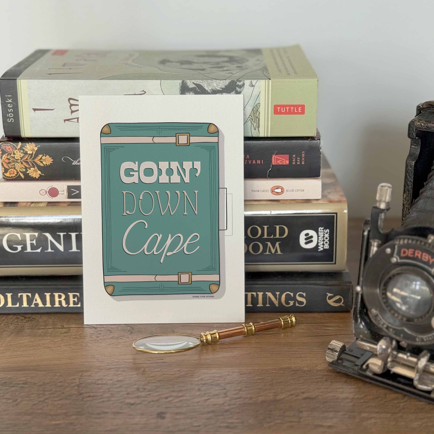 A notecard placed in front of a stack of books with a vintage camera and a small magnifying glass placed nearby. The artwork on the notecard is of a vintage style suitcase with the words "Goin Down Cape" drawn on it in unique lettering styles.